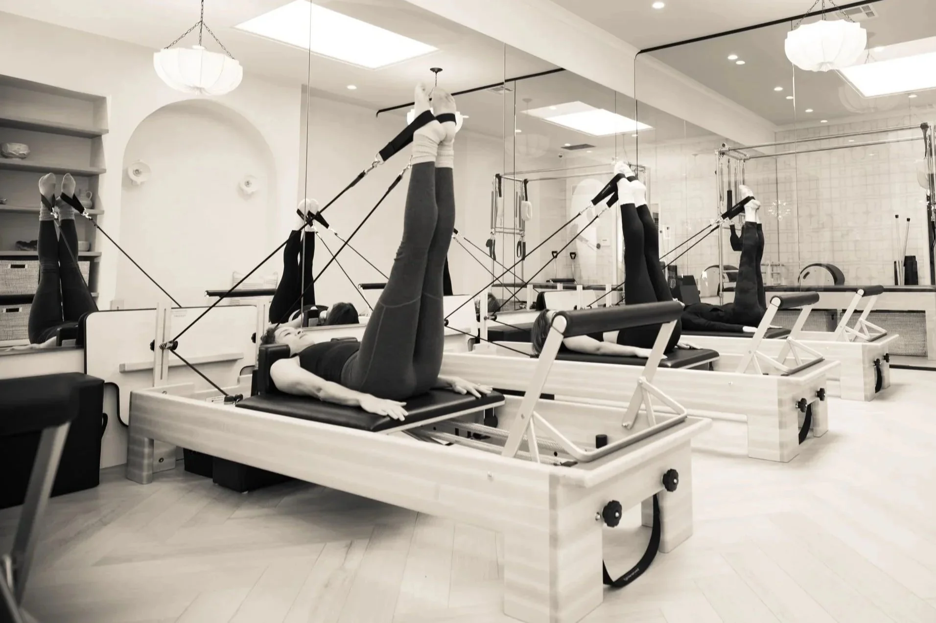 People exercising on Pilates reformers in a fitness studio with mirrors and bright lighting.