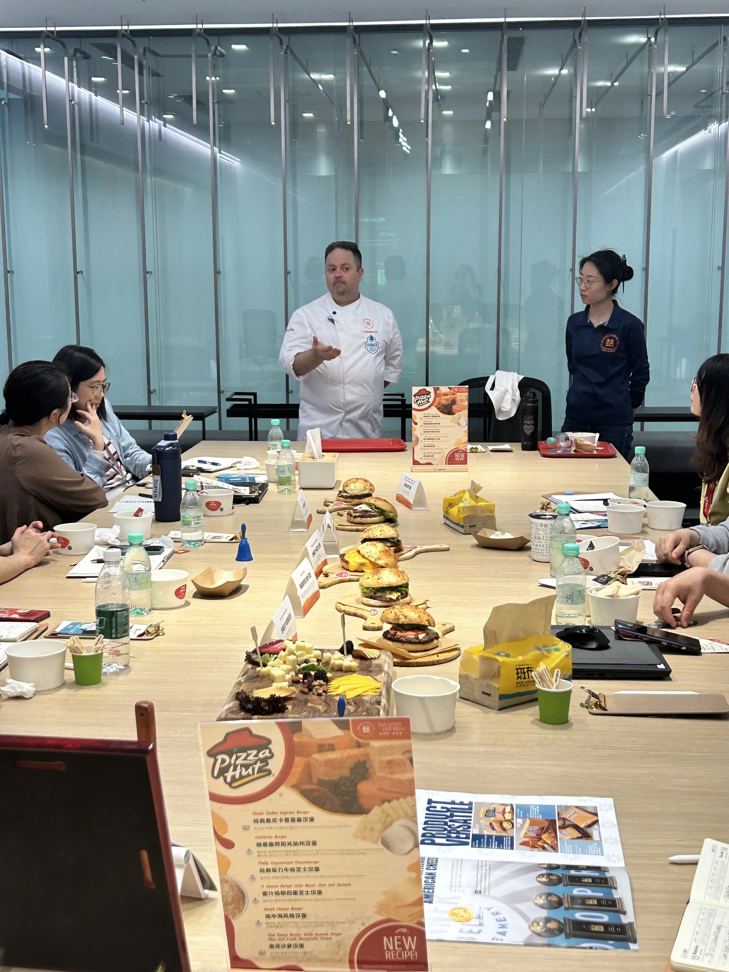 A group of people attending  food tasting or presentation in a conference room with a long table. The table displays various sandwiches, cheeses, and snacks, with food labels. A chef and an assistant stand at the head of the table talking to the group.