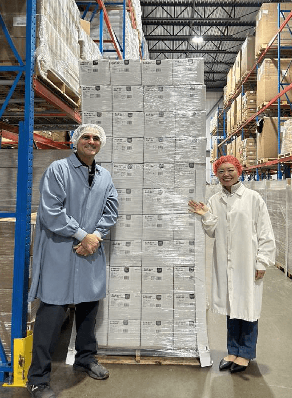 Two workers in protective clothing and hairnets standing next to a large pallet of boxed cheese products in a warehouse.