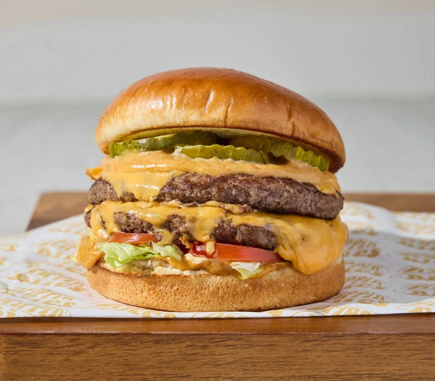 Close-up of a cheeseburger on a wooden surface, with pickles, lettuce, tomato, melted cheese, and two beef patties in a bun