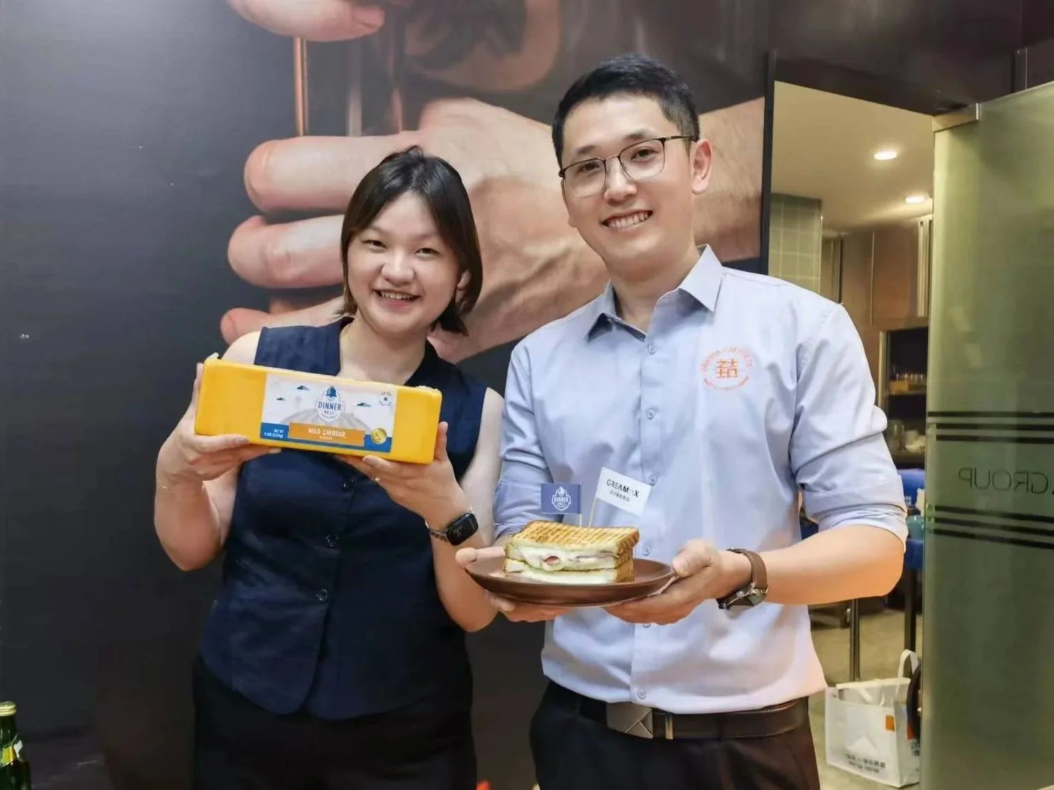 Two people smiling at a celebration; woman holding a yellow box labeled 'MILK CREAMER' and man holding a plate with a sandwich.