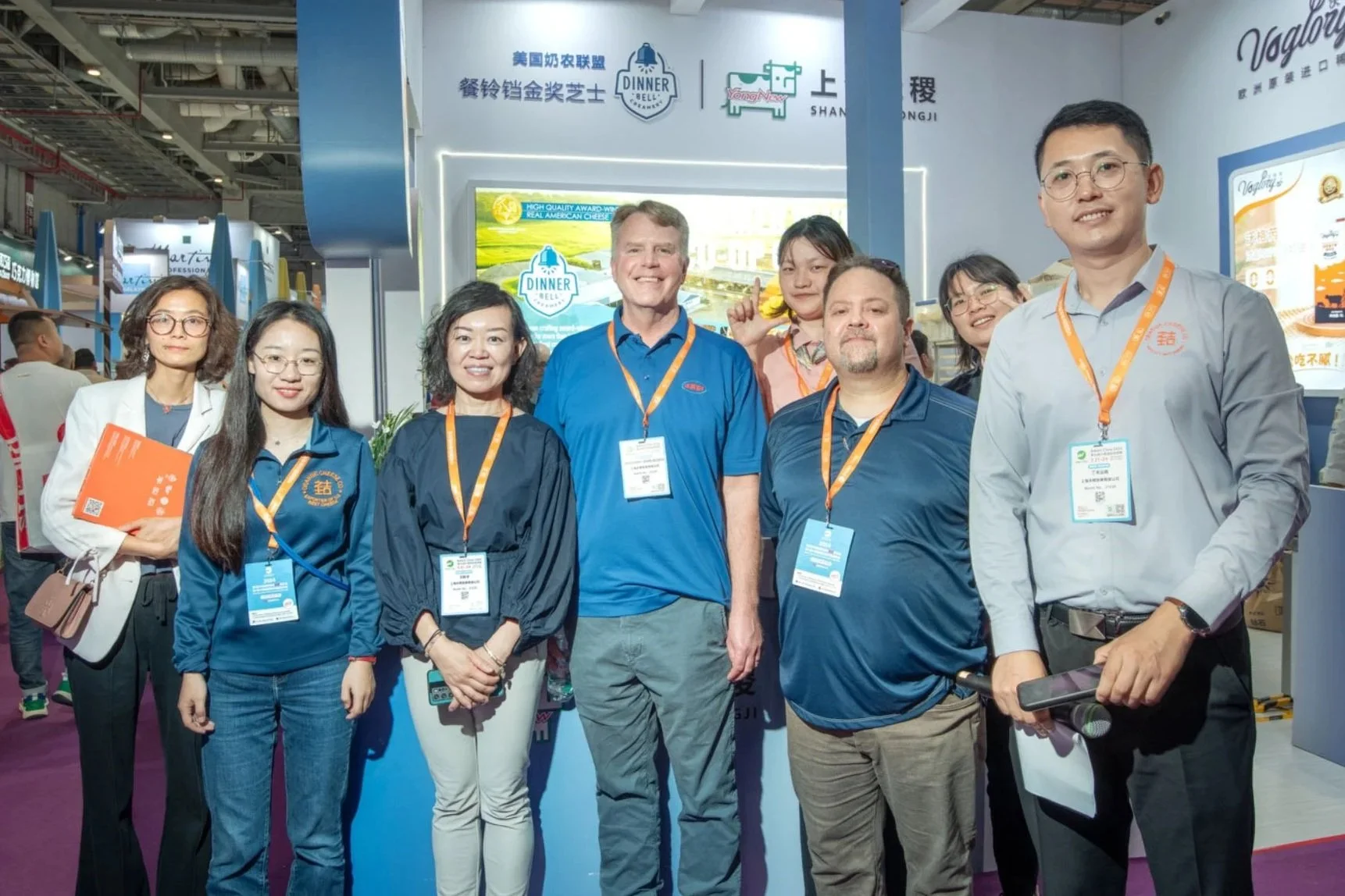 Group of nine people, including men and women of diverse ethnicities, posing together at a conference or trade show, with booths and signage in the background.