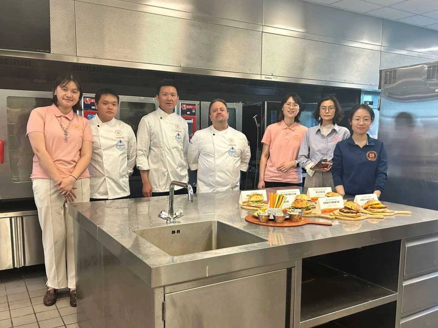 Group of seven people, including chefs in white coats and others in casual attire, standing behind a stainless steel kitchen counter with various sandwiches and food items displayed in front of them.