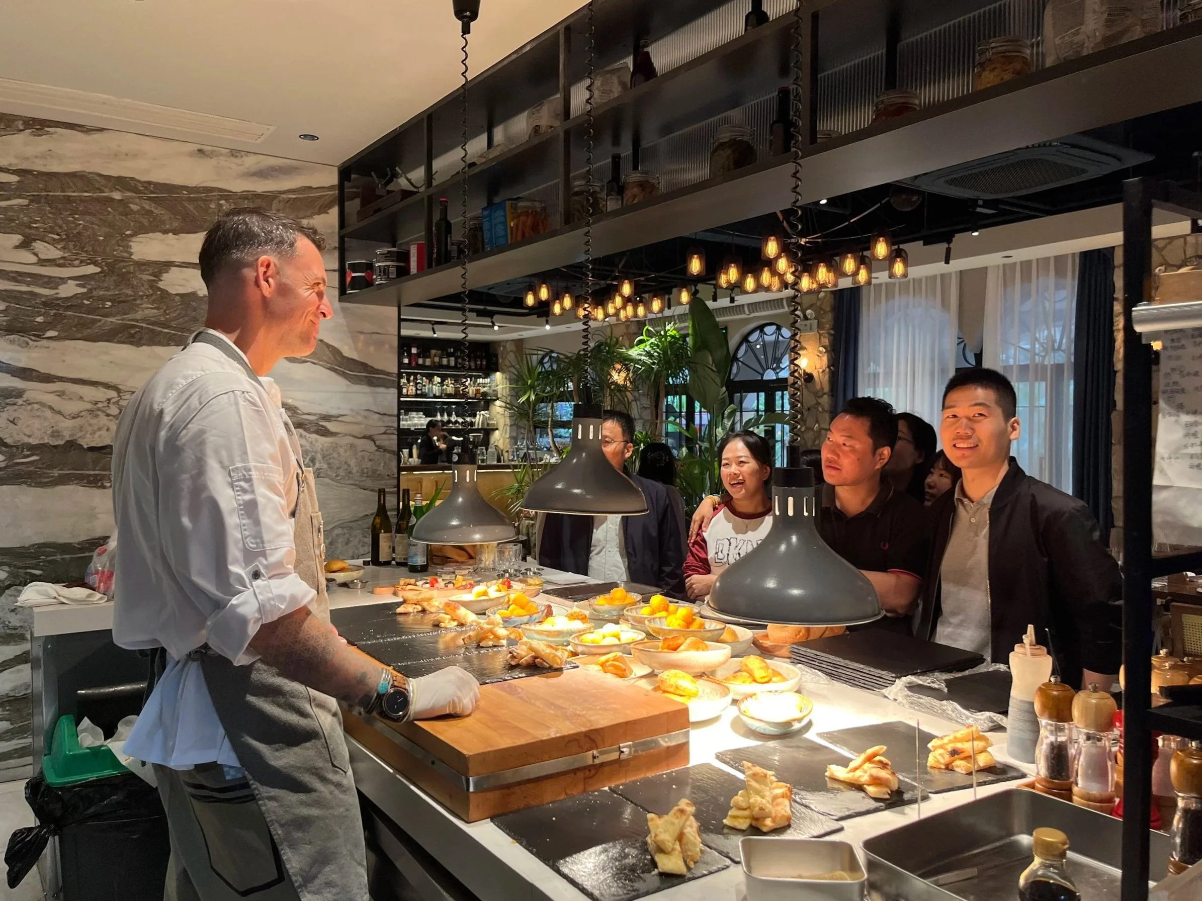 A chef presenting dishes at a restaurant counter with a group of smiling customers watching.