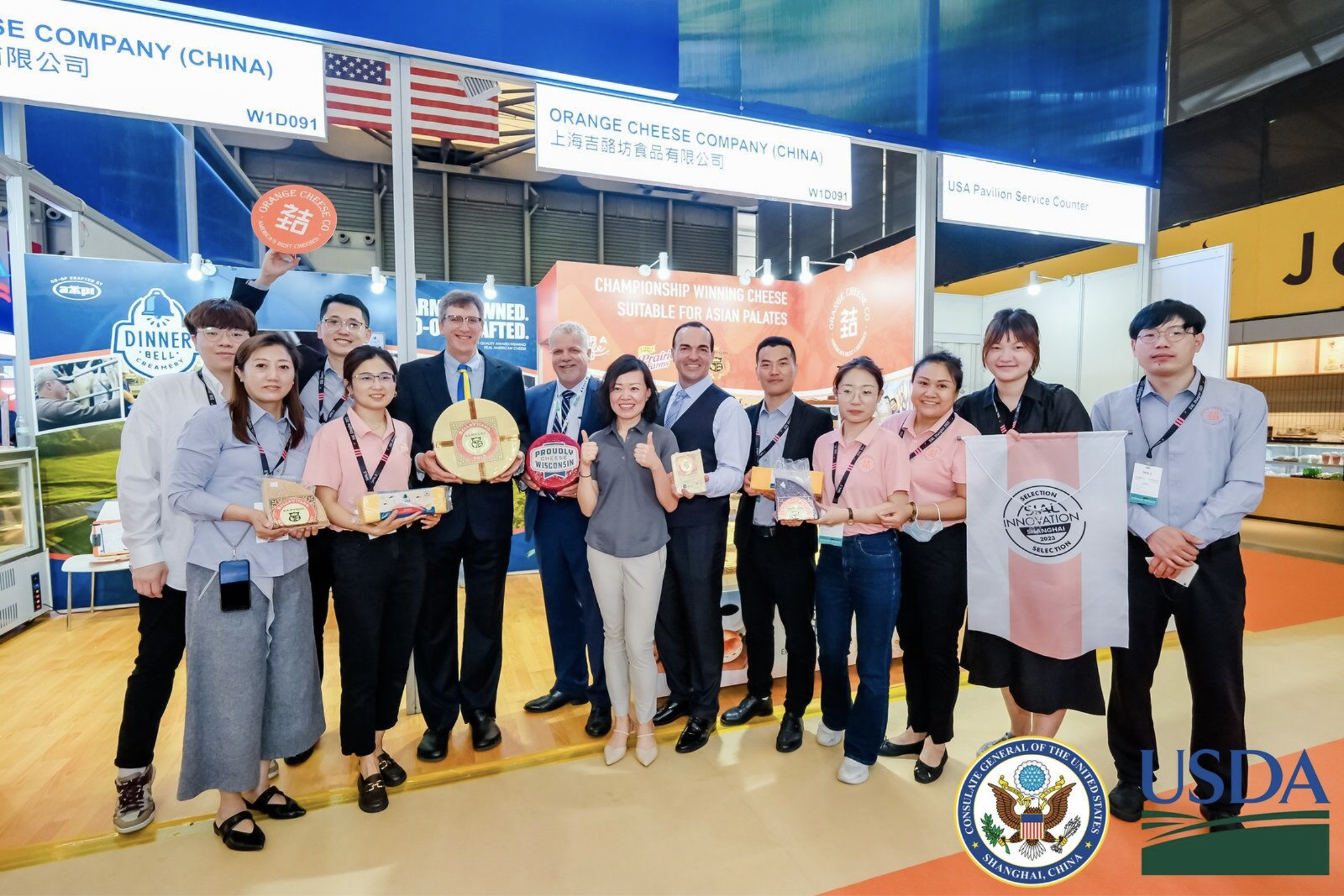 Group of people at an international food trade show, holding cheese and dairy products, with exhibitor booths and banners in the background.