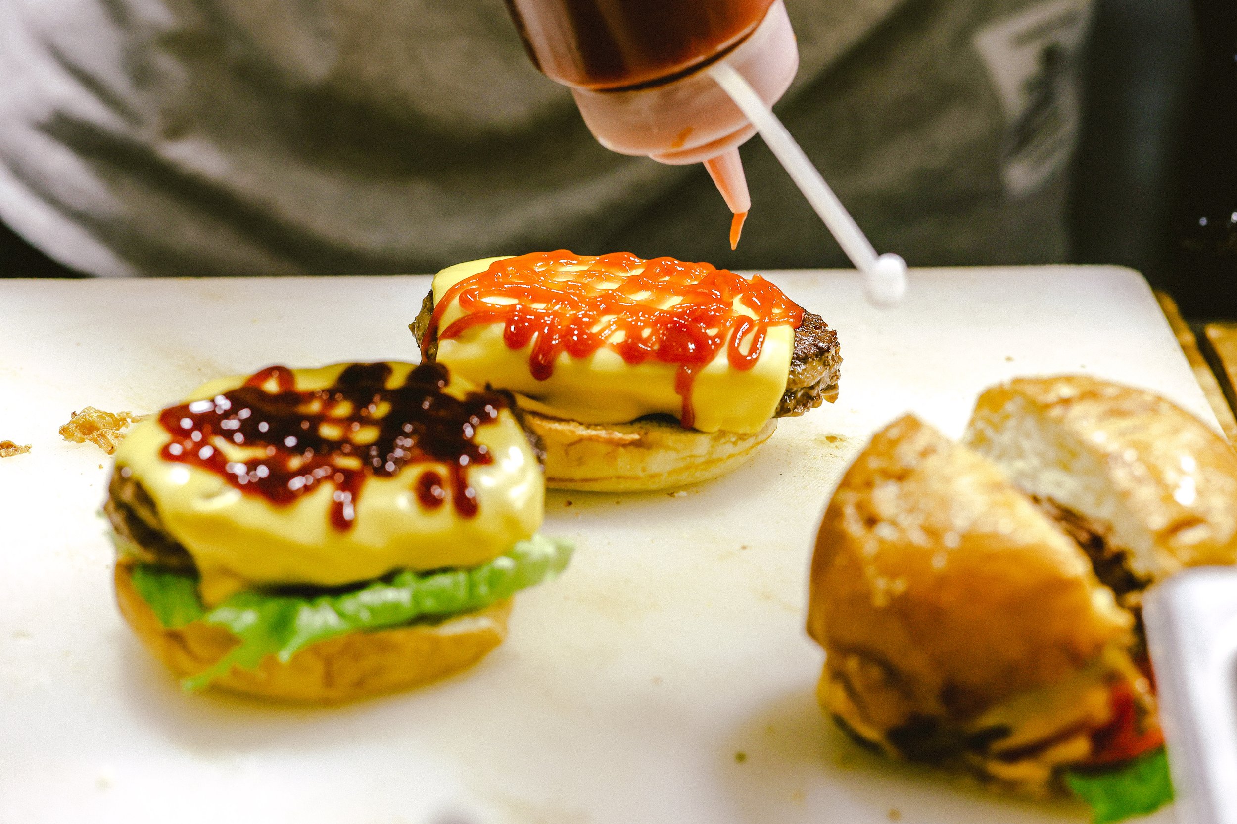 Close-up of a foodservice chef adding ketchup to two cheeseburgers on a white surface, with a fried chicken sandwich nearby.