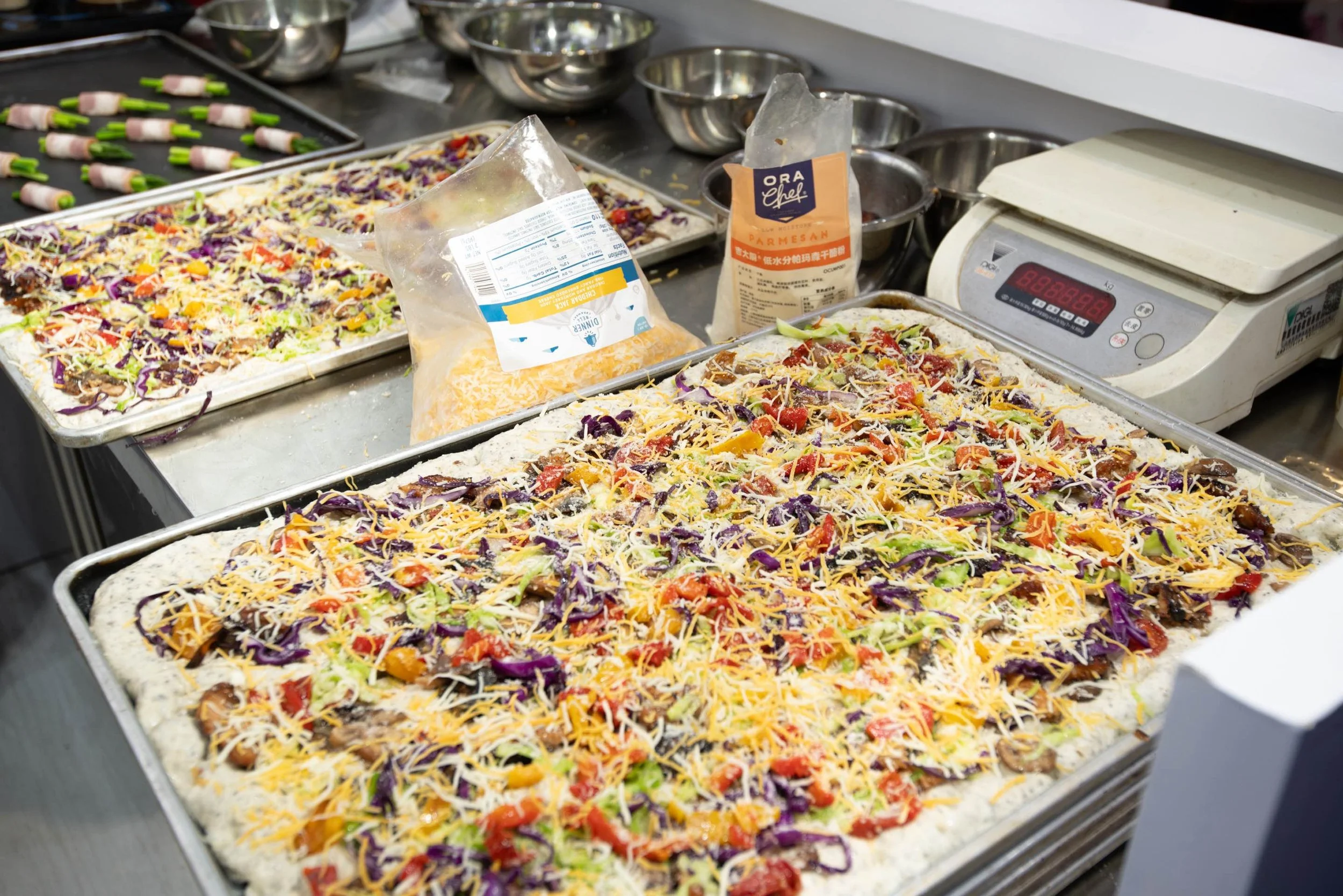 Unbaked pizza with shredded cheese, chopped vegetables, and sauce on a baking tray in a kitchen.