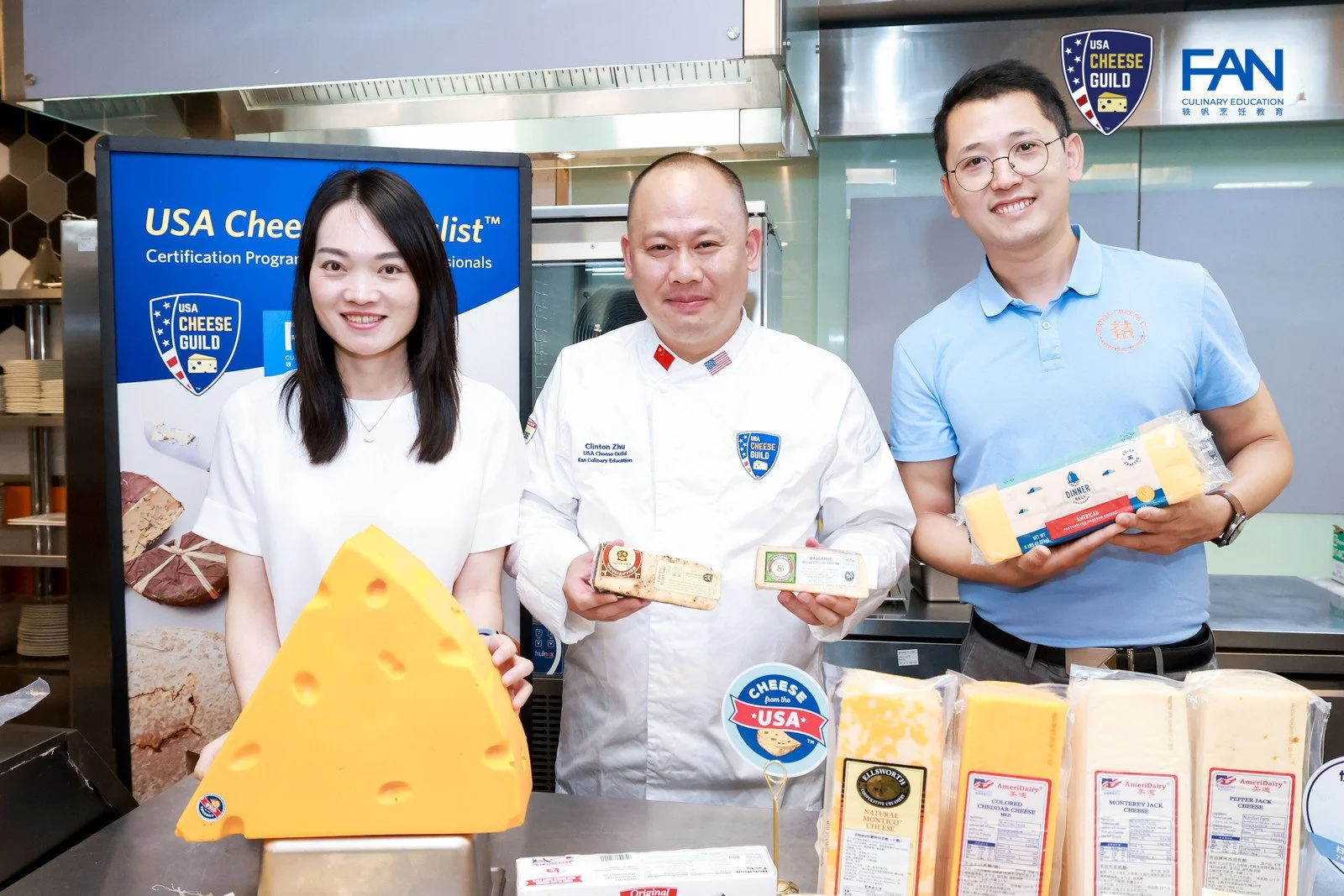 Three people standing behind a table displaying various cheeses, with a blue sign reading 'USA Cheese Guild' in the background. The person in the middle is wearing a white chef's coat, and the others are holding different cheese products.
