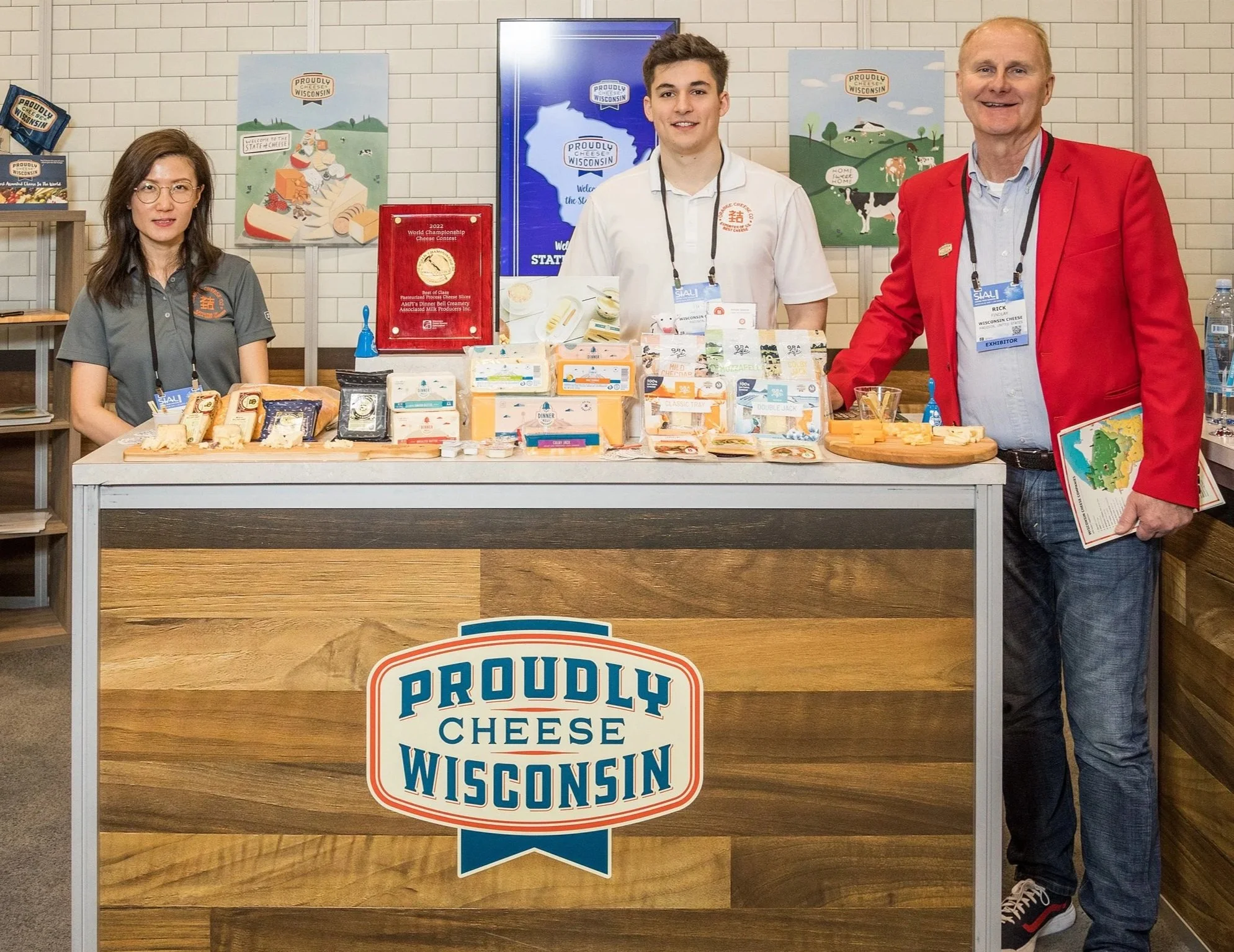 People standing behind a cheese display at the Proudly Cheese Wisconsin booth, with cheese varieties and promotional materials on the table.