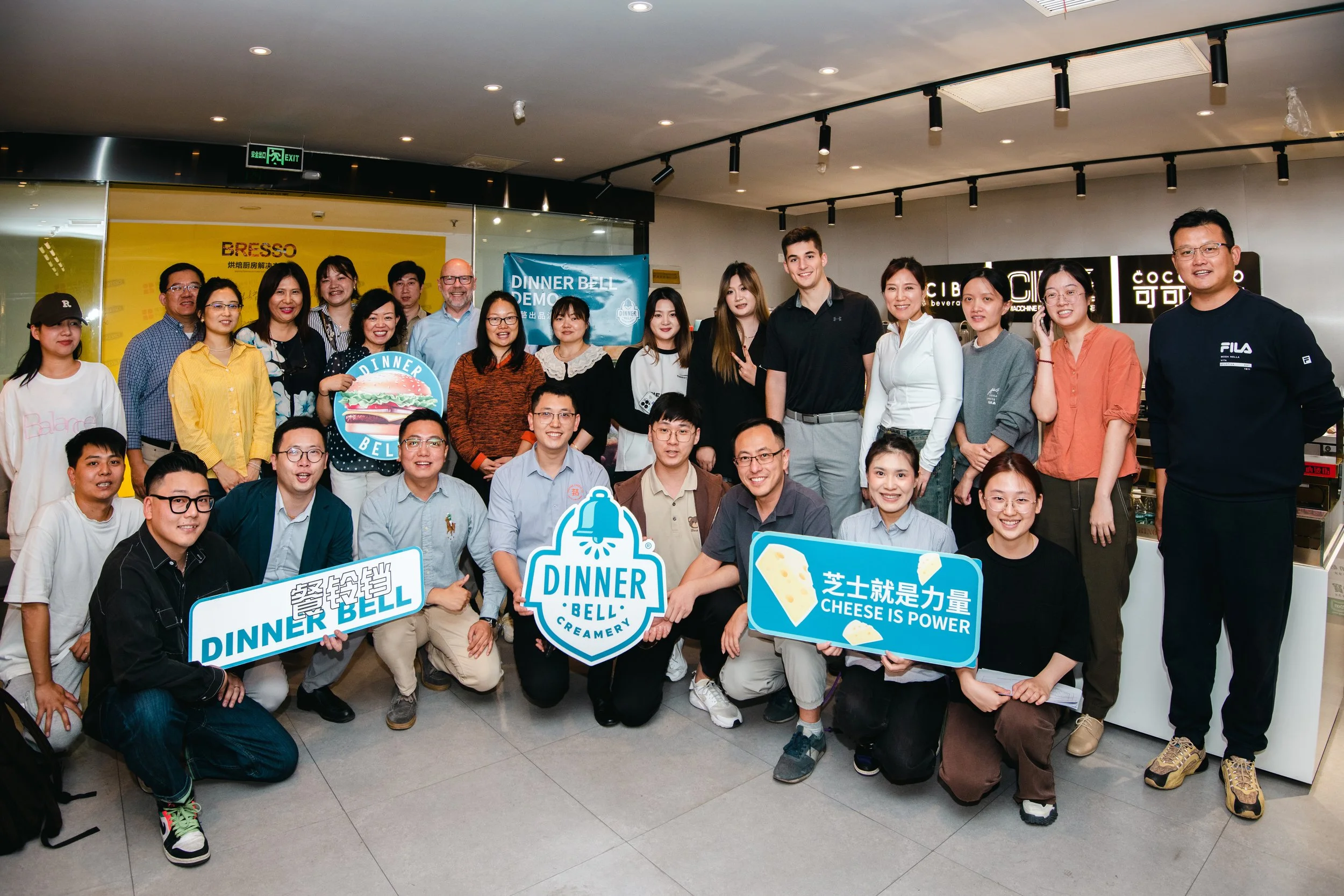 Group of people gathered in a restaurant or cafe, holding signs with slogans like 'Dinner Bell', 'Cheese is Power', and 'Dinner Bell Creamery'.