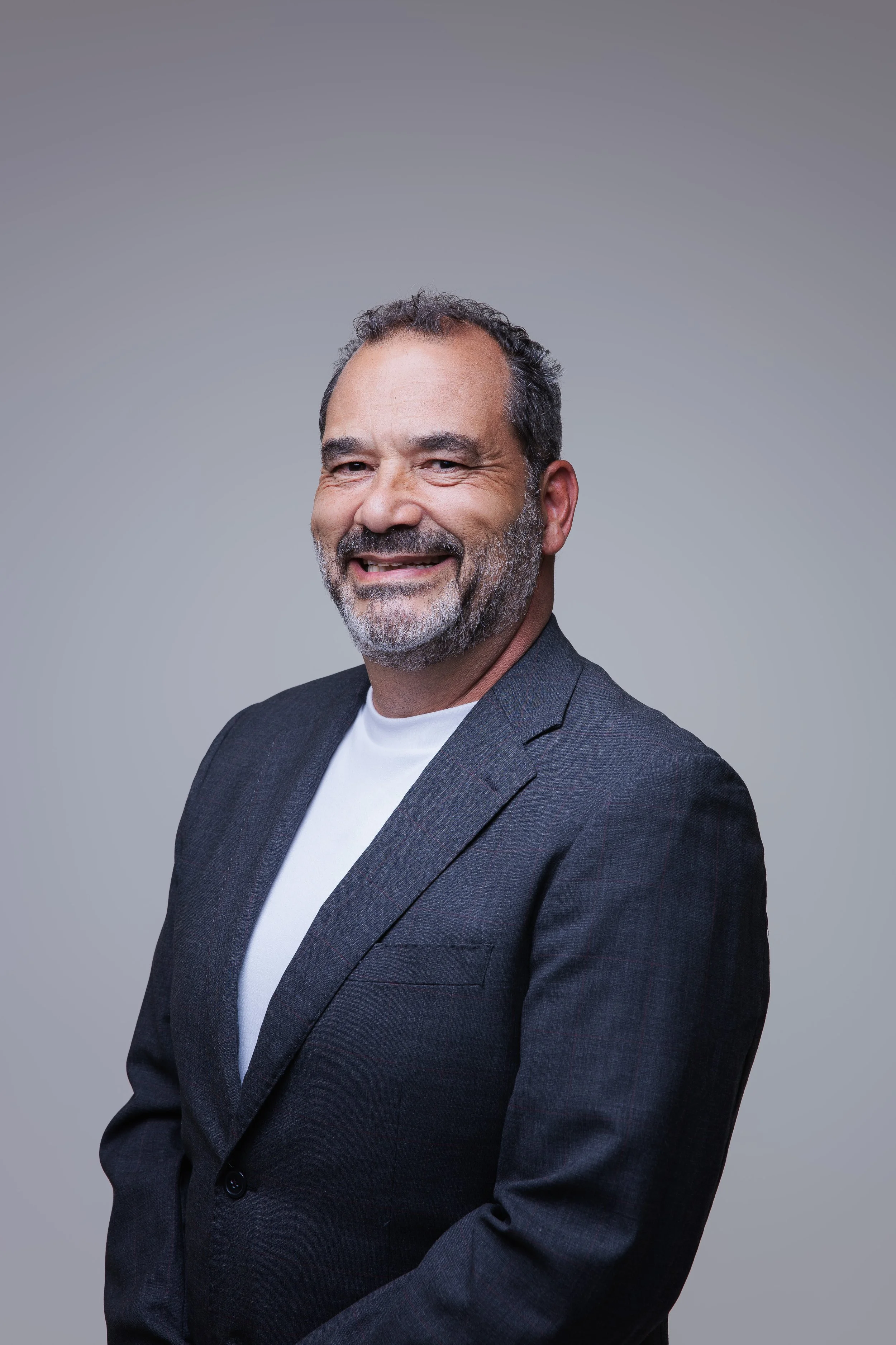 Homem sorridente com barba grisalha, cabelo curto, usando terno cinza e camiseta branca, posando contra fundo cinza