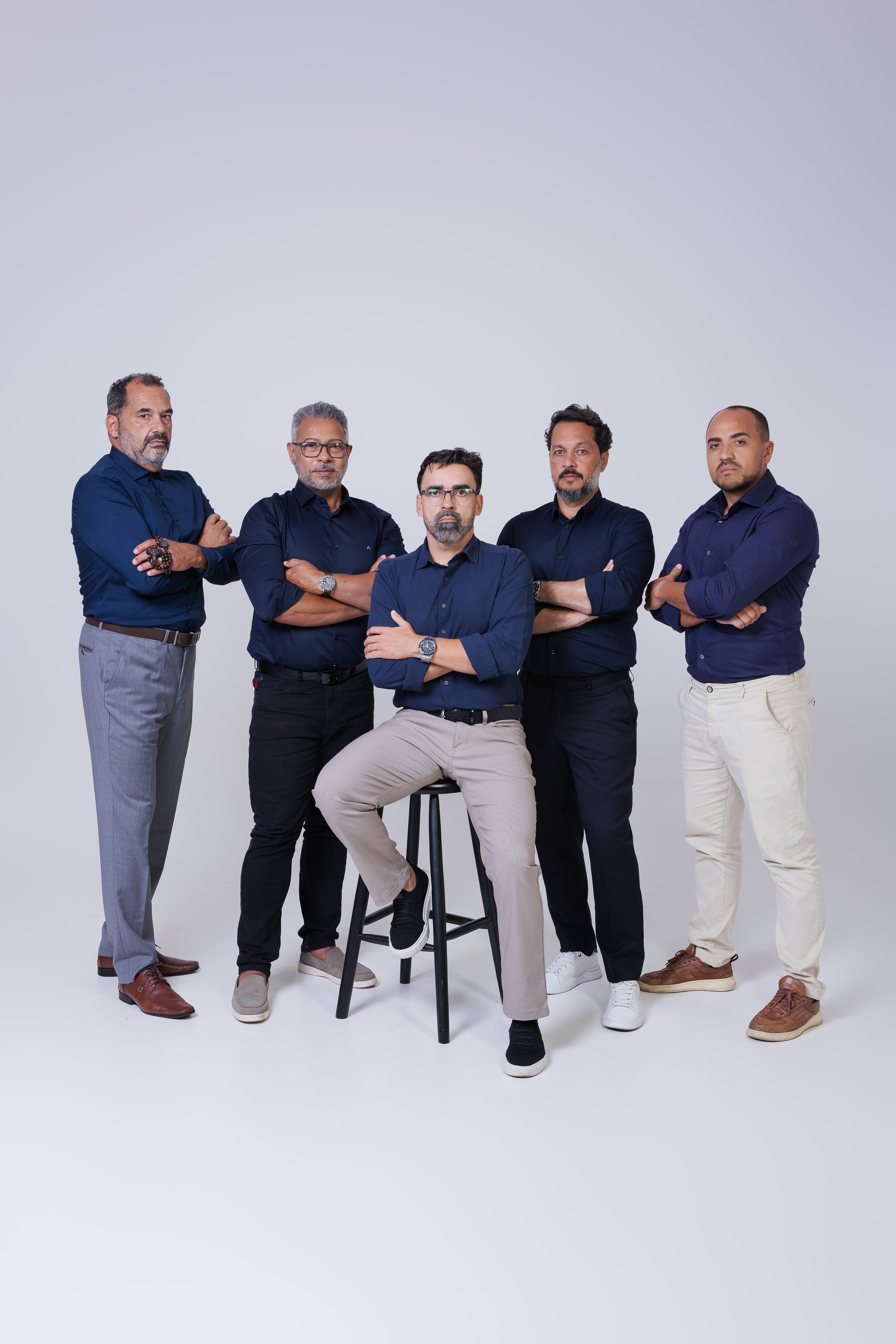 Grupo de cinco homens adultos com roupas casuals em uma parede branca, alguns com braços cruzados, um sentado em banquinho.