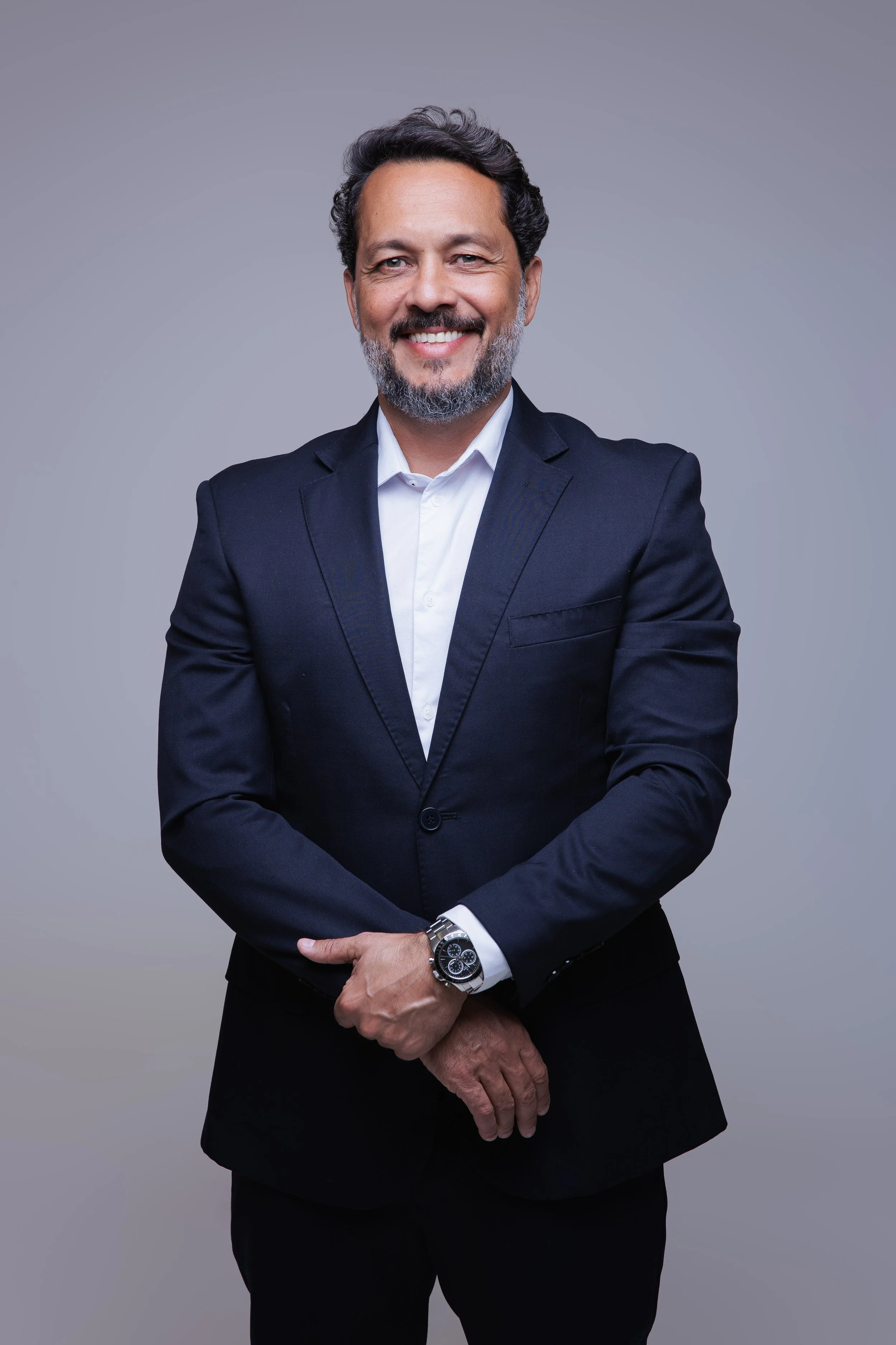 Homem sorridente usando terno escuro, branco, camisa com relógio no pulso, de cabelo e barba grisalhos, posando contra fundo cinza.