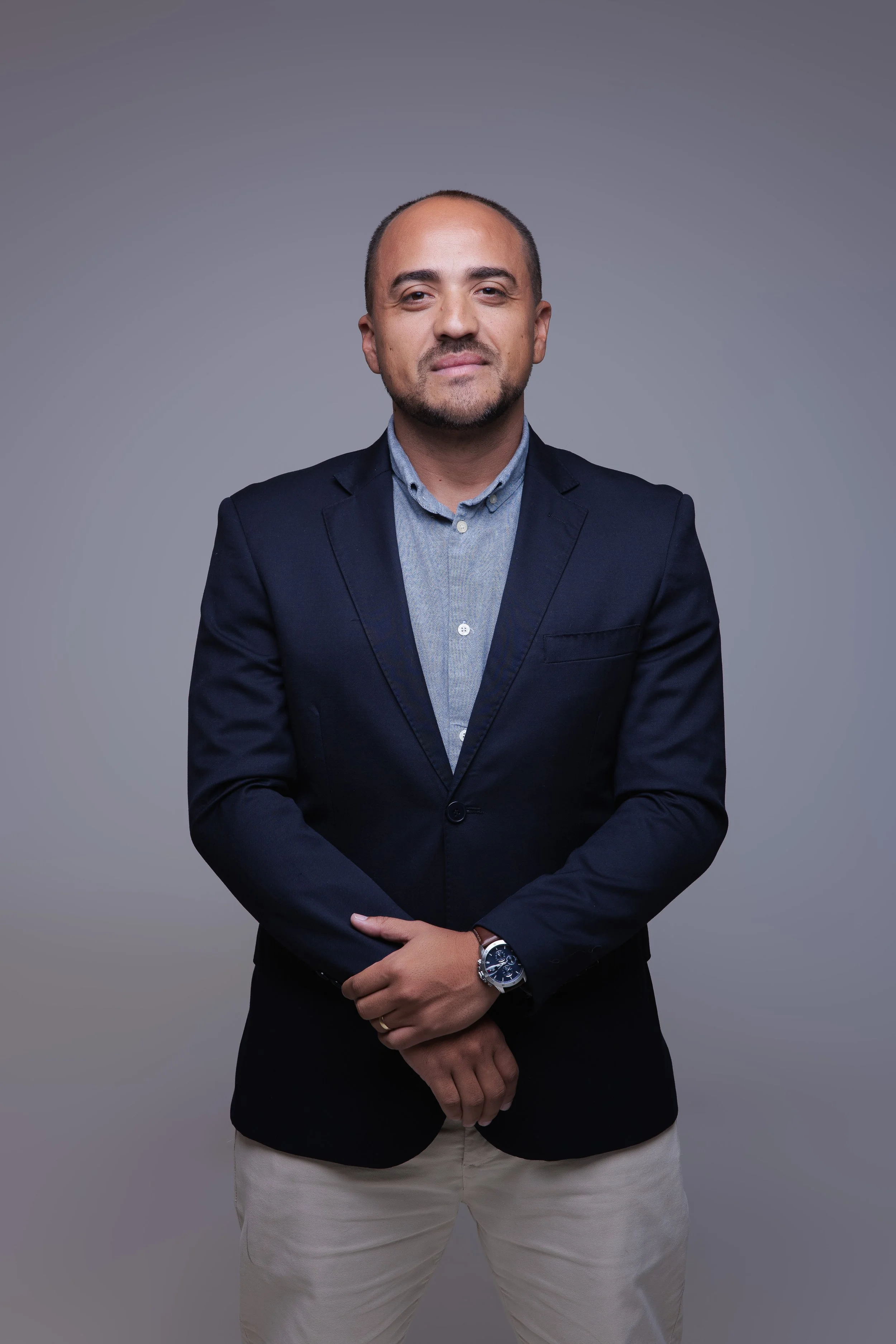 Homem vestindo blusa social, blazer navy, e calça bege, posando na frente de fundo cinza.