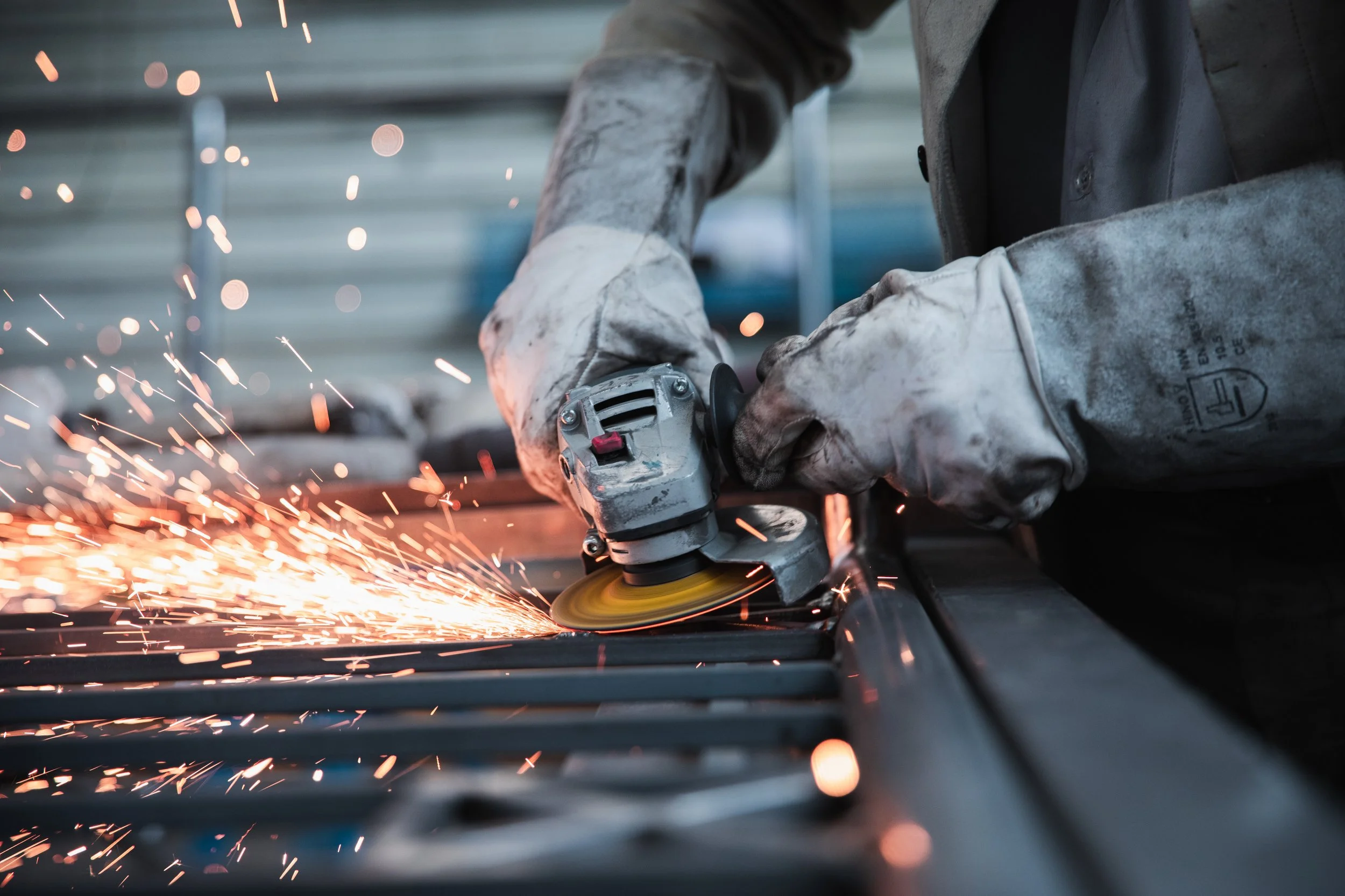 Pessoa usando óculos de proteção e luvas de trabalho, utilizando uma ferramenta de usinagem para cortar metal, com faíscas voando.