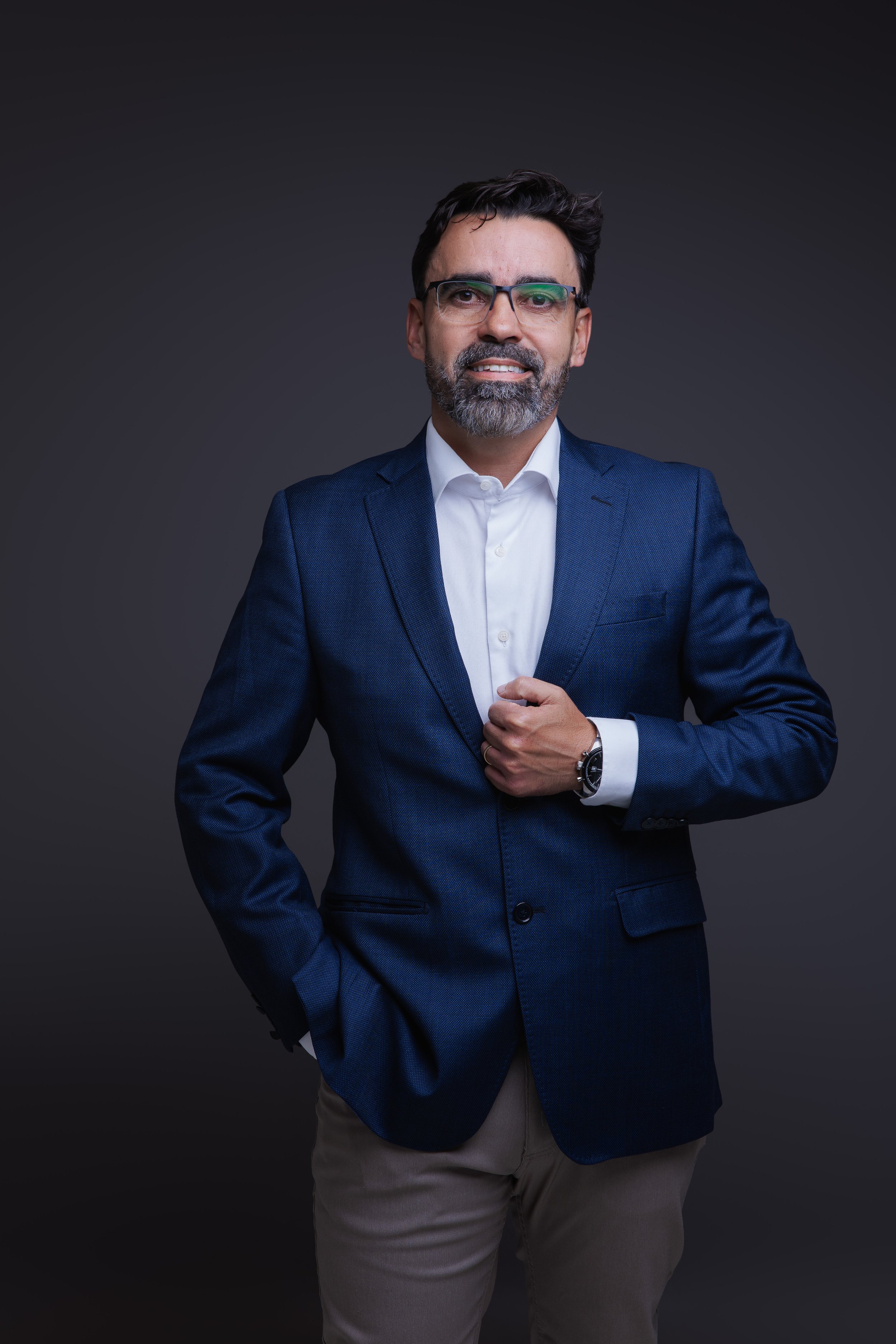Homem de meia-idade, vestindo terno azul, camisa branca, com cabelo escuro, barba e óculos, posando contra fundo escuro