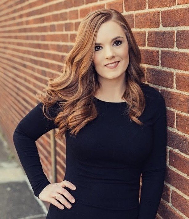 Young woman with long, wavy auburn hair leaning against a red brick wall, wearing a black long-sleeve top.