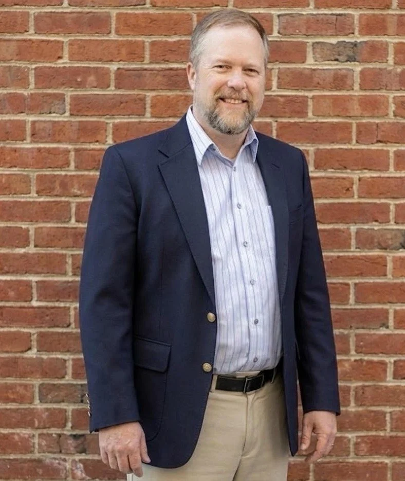 A middle-aged man with a beard and short hair wearing a navy blazer, light blue striped shirts, and beige pants, standing in front of a red brick wall, smiling at the camera.