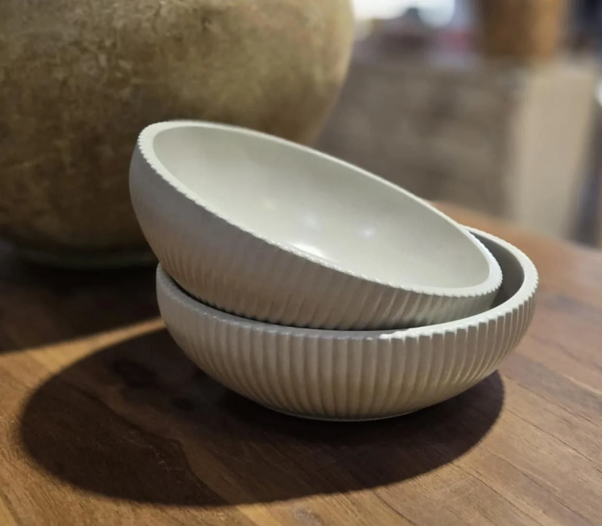 Two white ceramic bowls with ribbed exterior stacked on a wooden surface, with a large beige ceramic pot in the background.