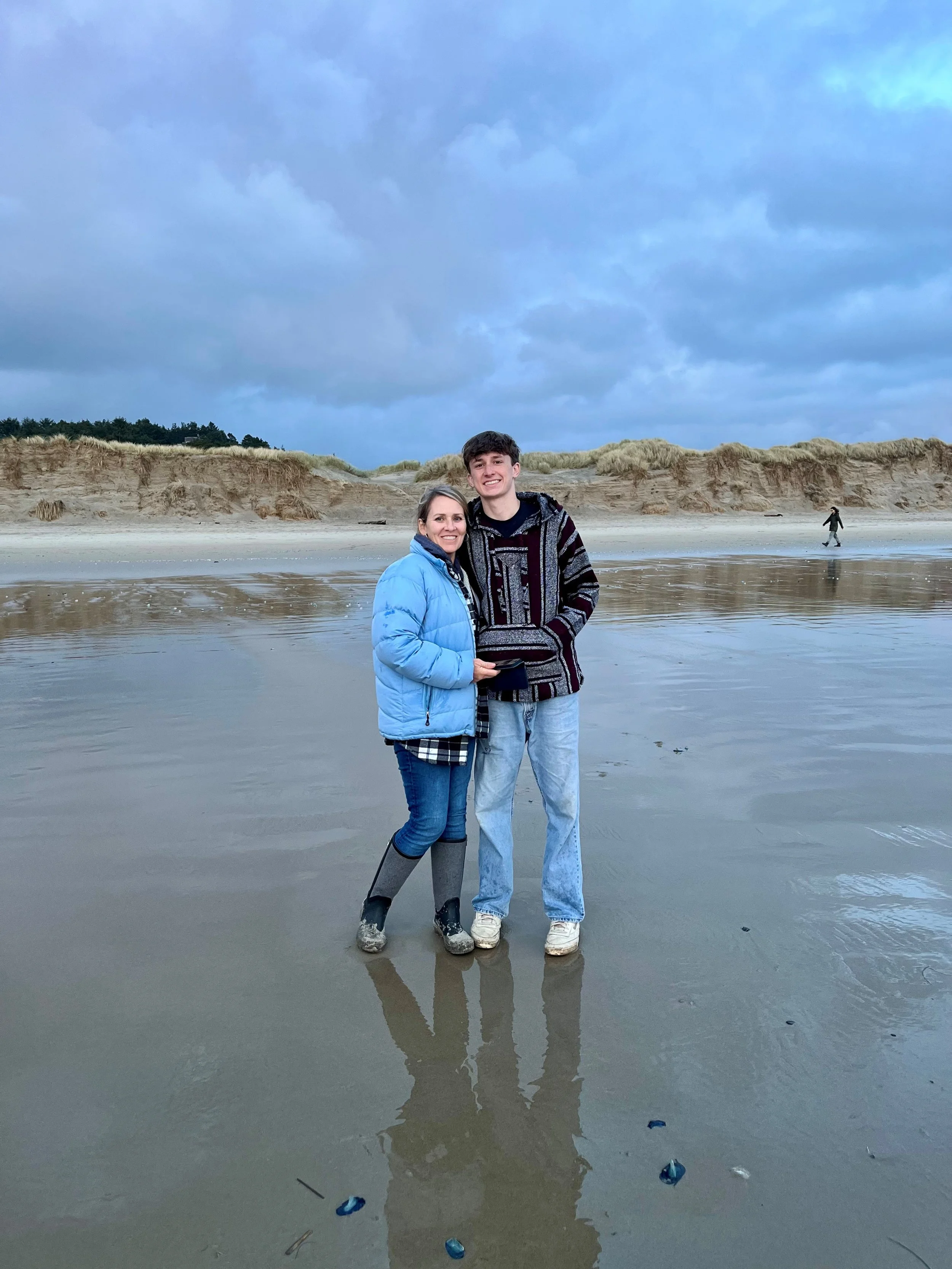 A woman and a young man standing on a wet beach, smiling at the camera. The woman is wearing a blue jacket and black rain boots, and the young man is wearing a patterned sweater and jeans. There are sand dunes and some clouds in the sky in the background.