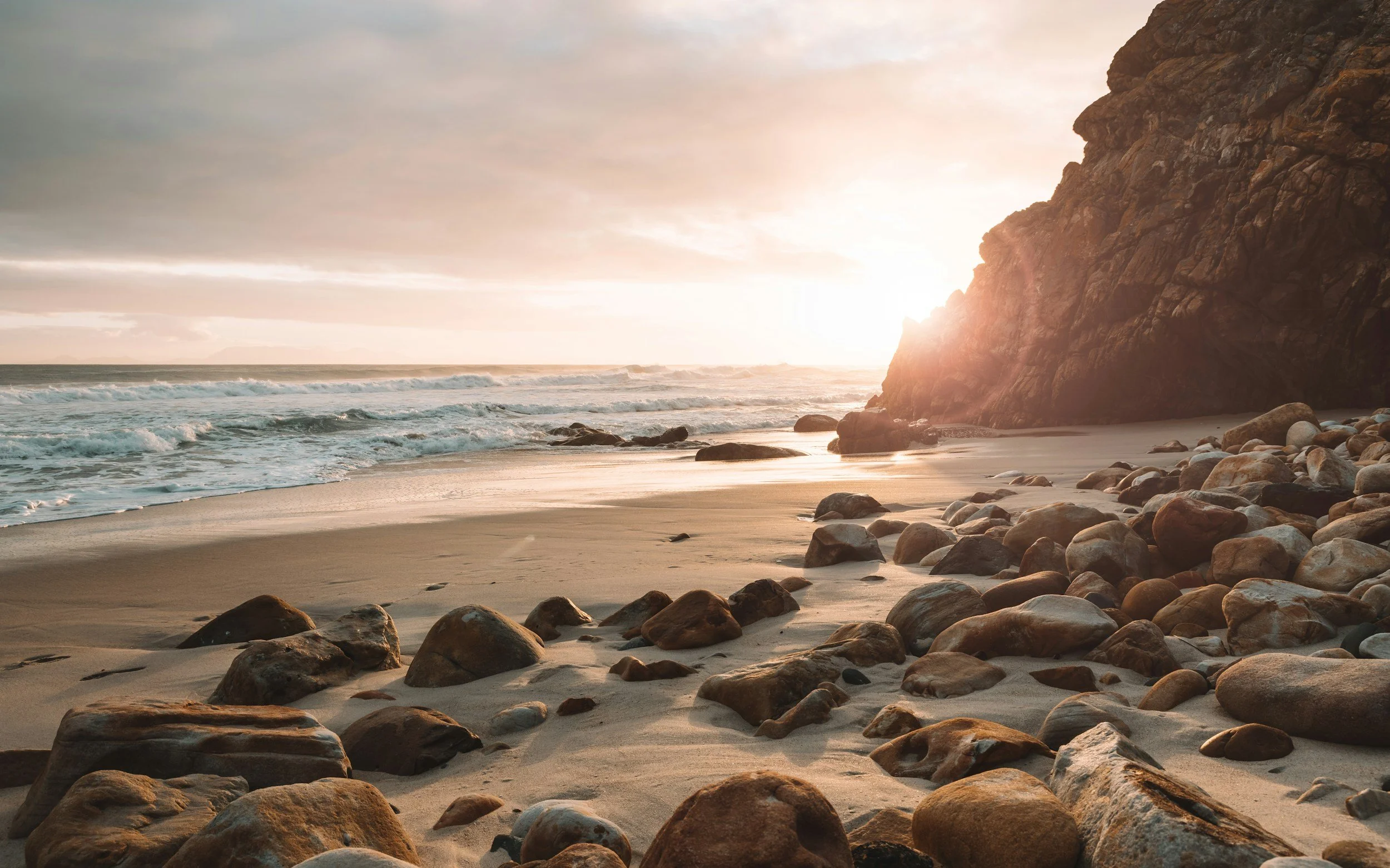 A rocky beach at sunset with large rocks scattered on the sandy shore, ocean waves, and a cliff on the right side.