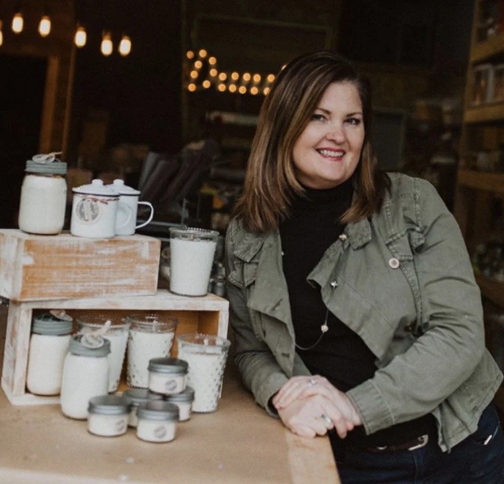 A woman with shoulder-length brown hair smiling and leaning on a wooden table with jars and containers of homemade products in a cozy, dimly lit shop or studio.