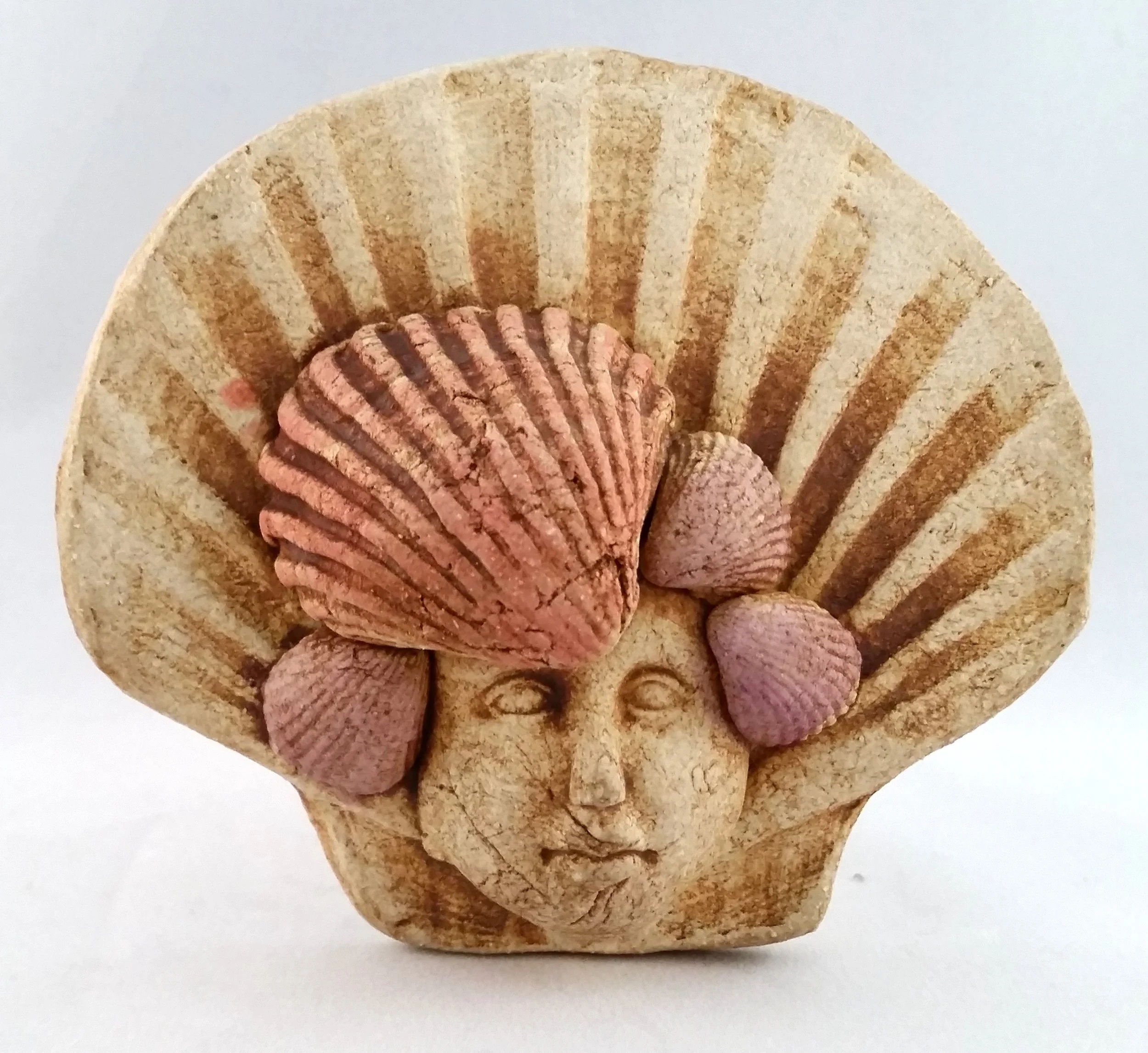 Decorative sculpture of a woman's face with a shell hairpiece and small shells on the sides, set against a plain background.
