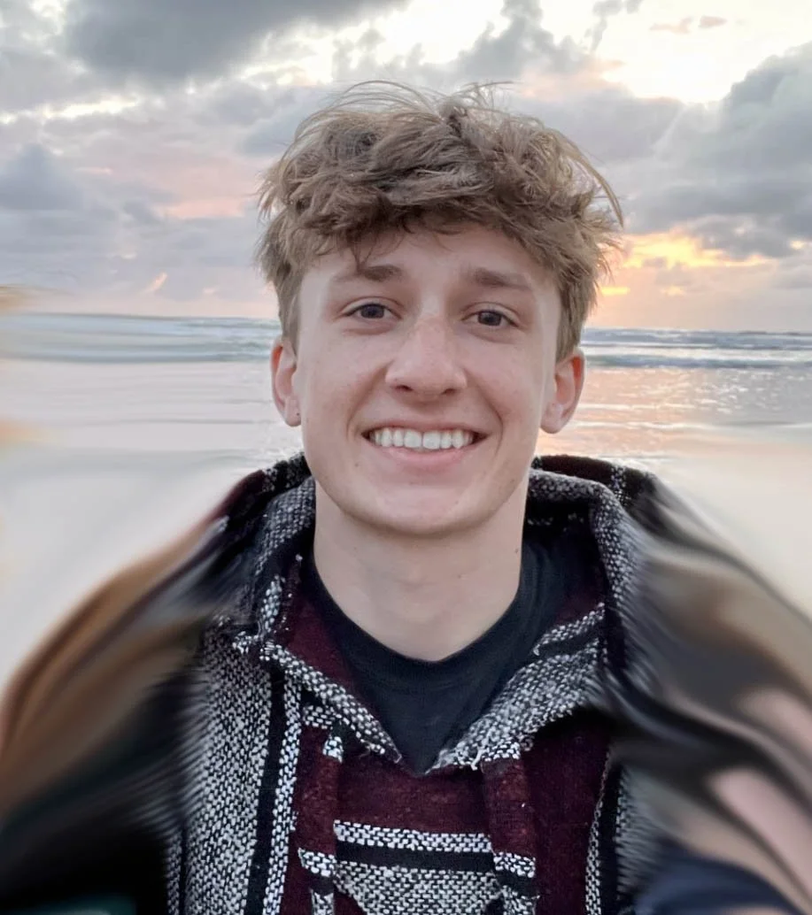Young man taking a selfie on the beach during sunset, with ocean waves and cloudy sky in the background.