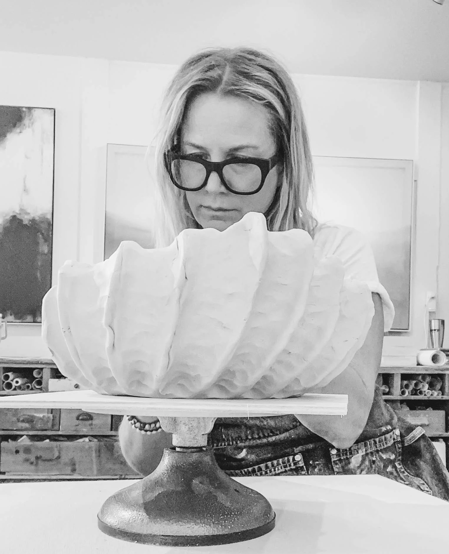 A woman with long hair, wearing glasses, is working on a carved shell-shaped object on a pedestal in a room that appears to be an art studio.