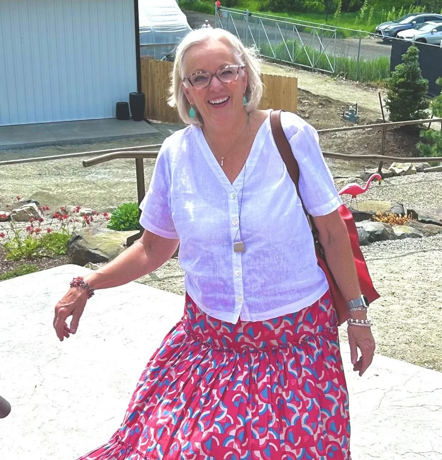 A woman with blond hair, glasses, and turquoise earrings smiling outdoors in front of a garden and a parking lot. She is wearing a white short-sleeved blouse, a colorful patterned skirt, and has a brown purse over her shoulder.