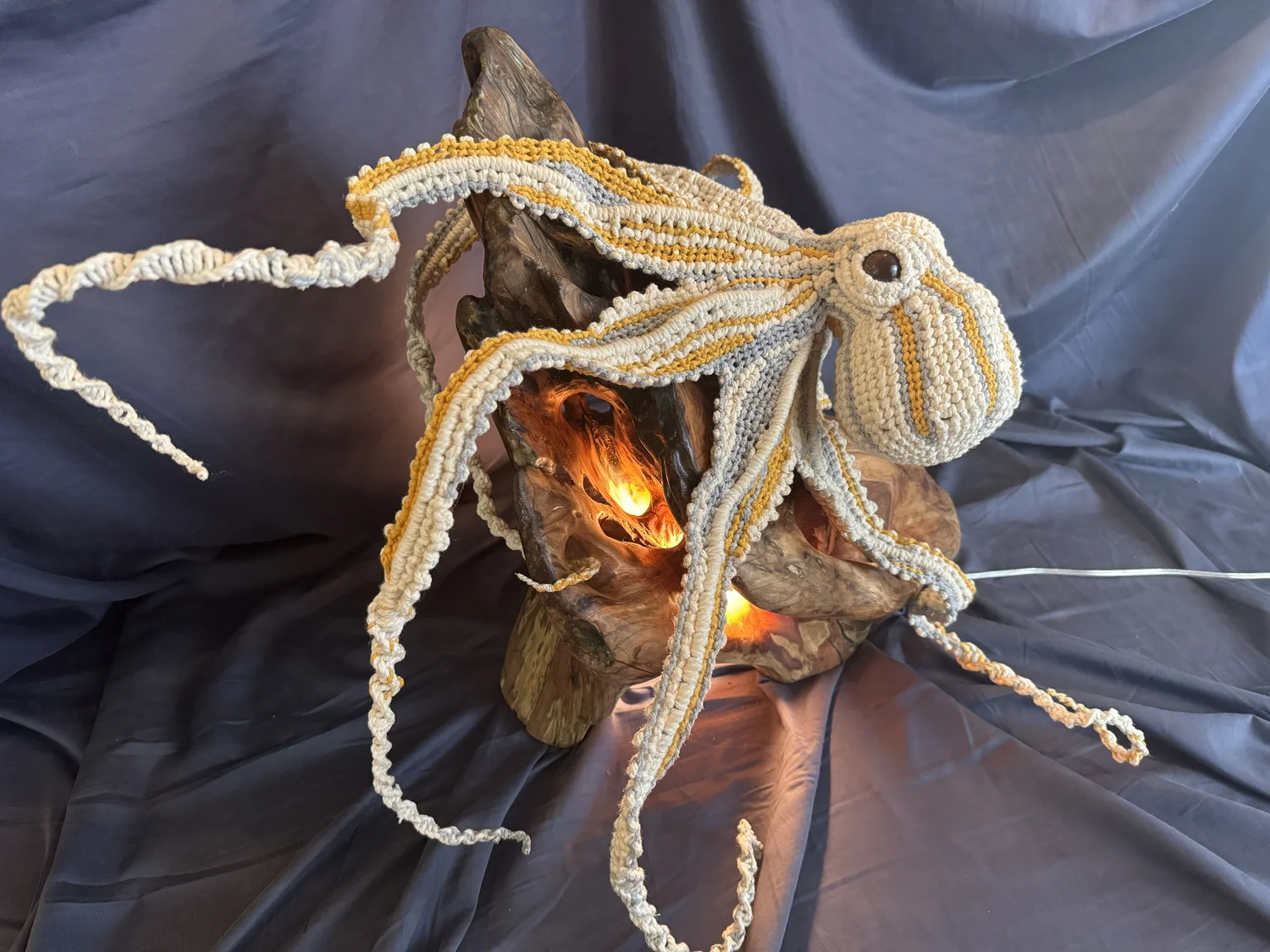 A knitted octopus with beige, white, and yellow yarn, draped over a piece of driftwood on a dark fabric background.