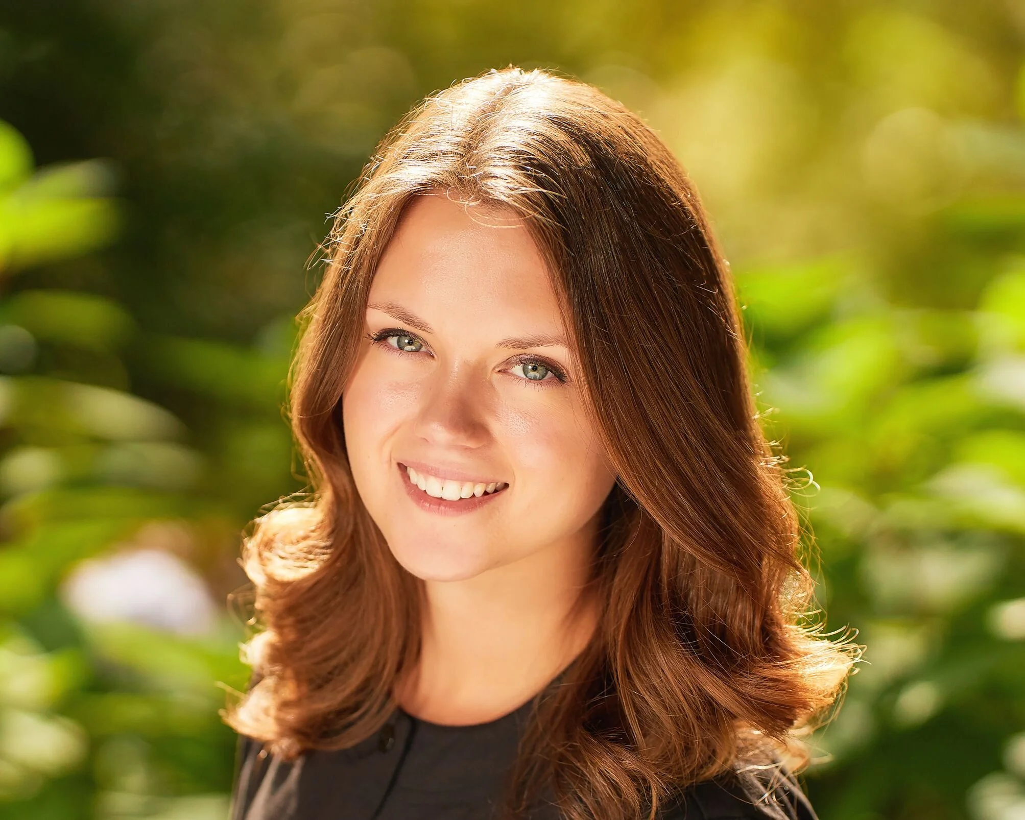 A woman with long, wavy brown hair and blue eyes smiling outdoors with blurred green foliage in the background, illuminated by warm sunlight.