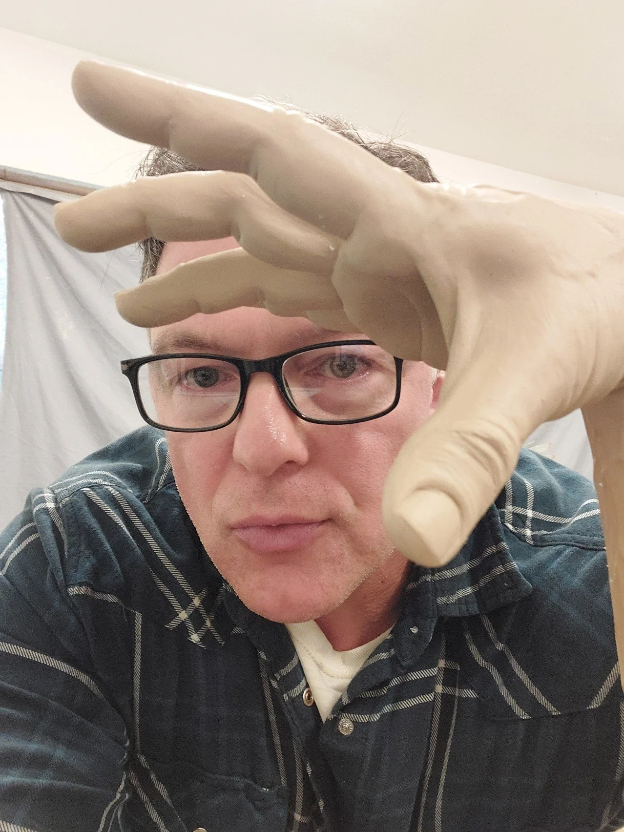 A person wearing glasses and a plaid shirt is reaching towards the camera with a sculpted hand, partially covering their face.