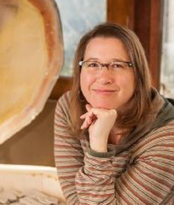 A woman with glasses and shoulder-length brown hair smiling, resting her chin on her hand.