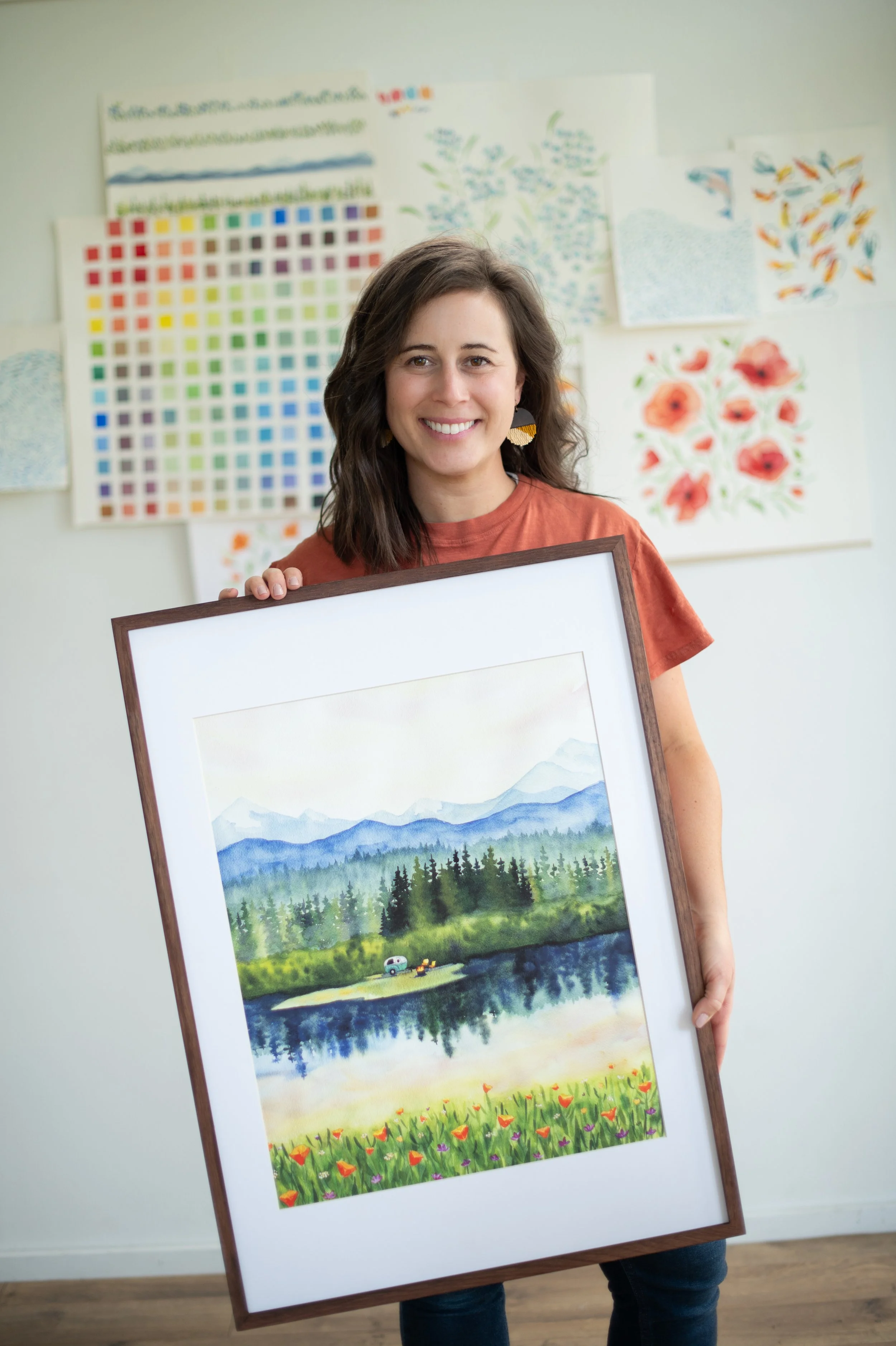 A woman smiling and holding a framed watercolor painting of a mountain landscape with lake, forest, and field of flowers, standing in an art studio with colorful artwork and swatches on the wall behind her.