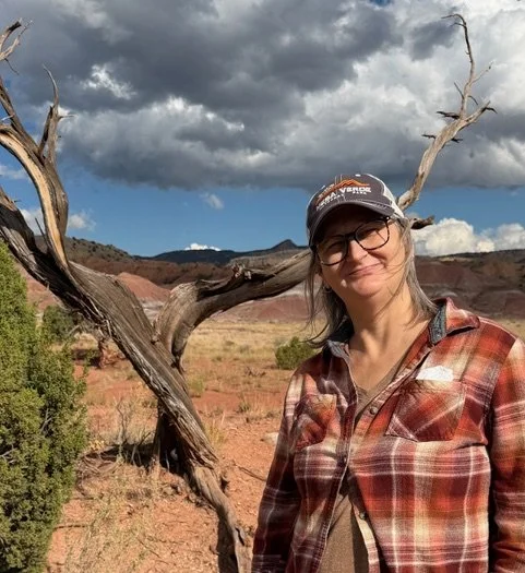 Woman wearing glasses, a cap, and a plaid shirt standing next to a leafless tree in a desert landscape with mountains and cloudy sky in the background.