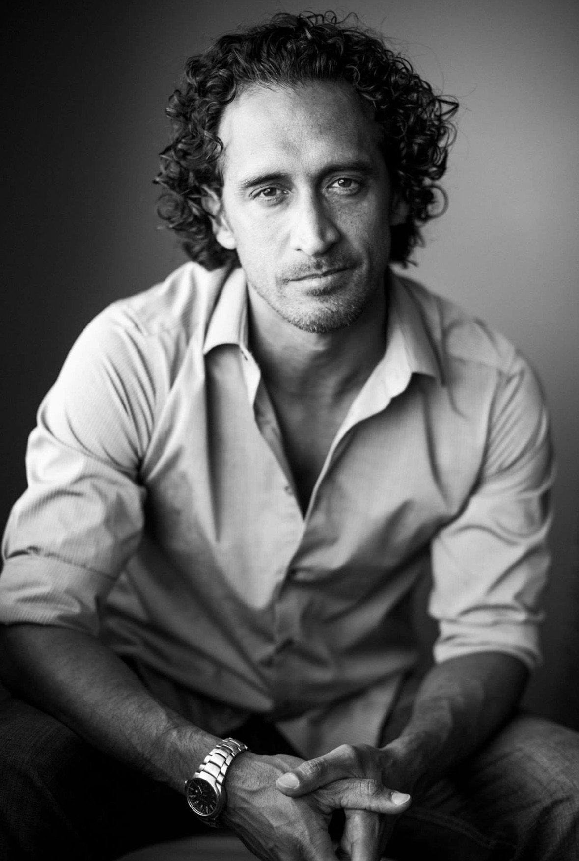 Black and white portrait of a man with curly hair, wearing a light-colored button-up shirt, sitting with his hands clasped, looking at the camera.