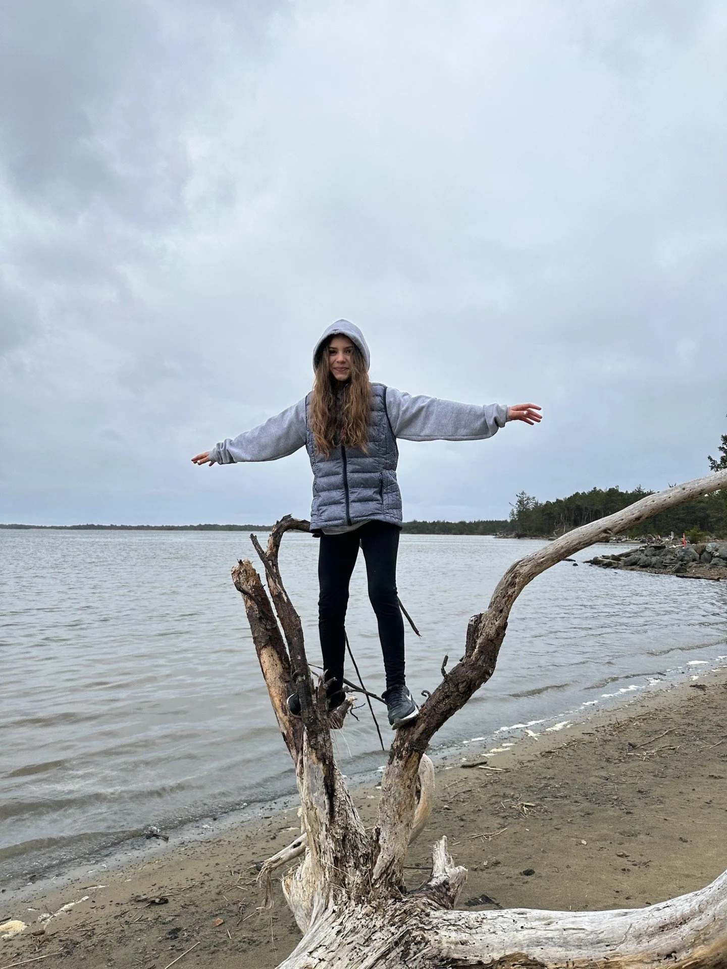 A young woman stands on a large, weathered tree branch at the beach with arms outstretched, wearing a gray hoodie, gray vest, and black pants, under a cloudy sky.