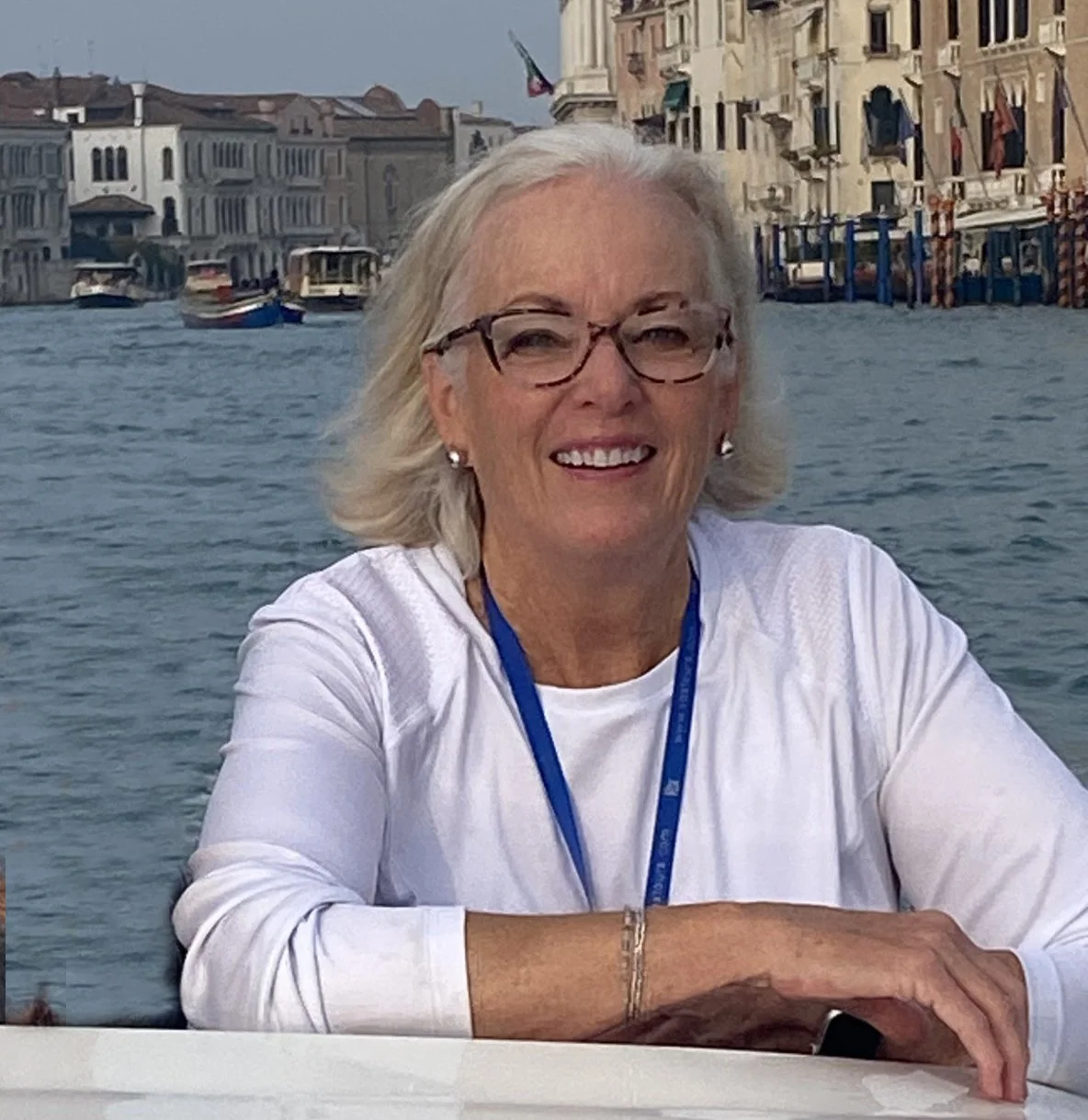 An woman with gray hair and glasses smiling, seated by a canal with boats and historic buildings in the background, during daytime.