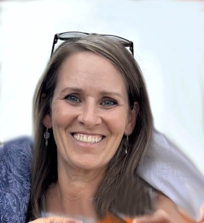 A woman with long brown hair, wearing earrings and glasses on her head, smiling at the camera.