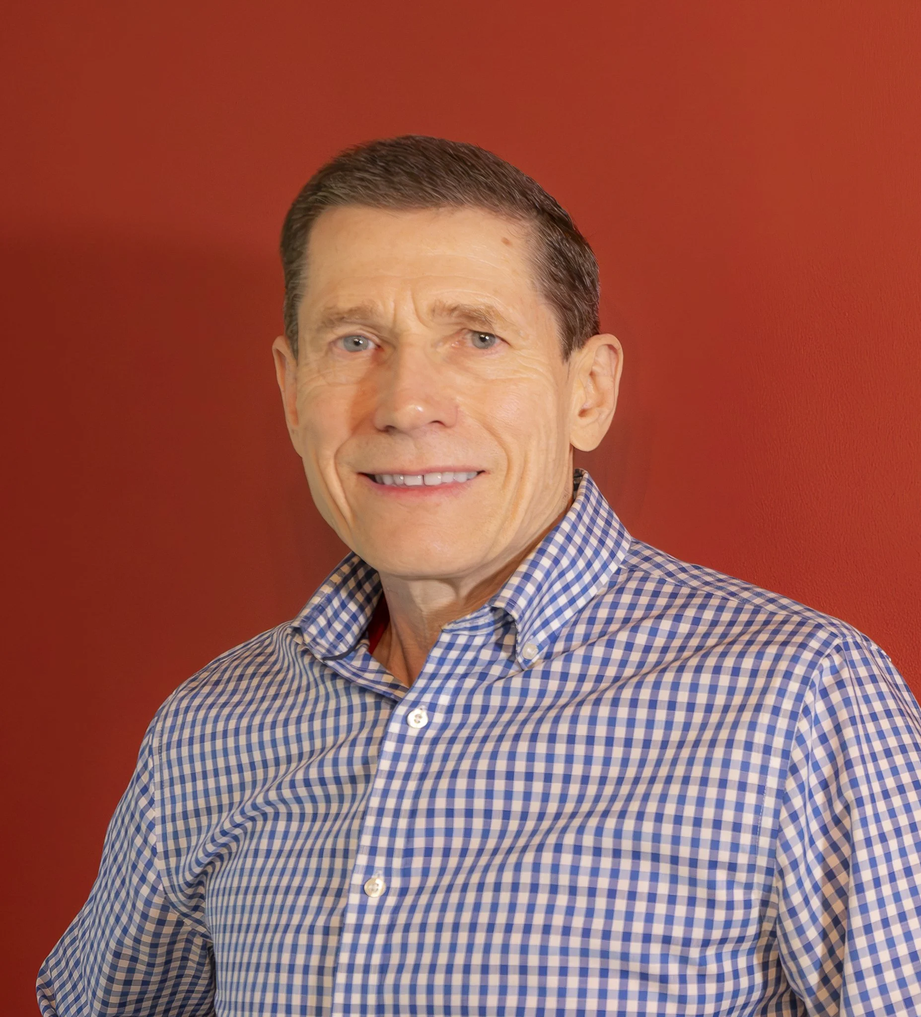 A middle-aged man with short brown hair, blue eyes, smiling, wearing a blue and white checkered button-up shirt, standing against a red background.