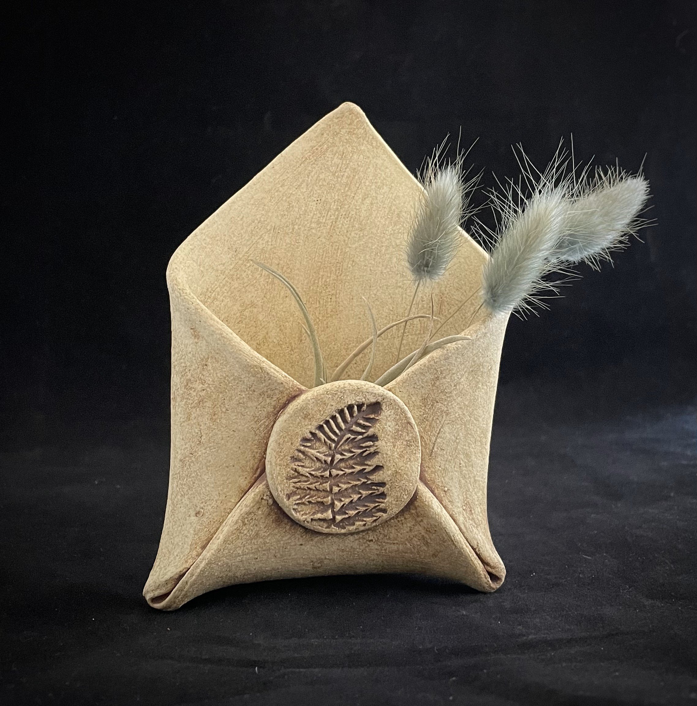 A beige velvet pouch with a circular seal featuring a fern leaf impression, holding three dried fuzzy gray and white grass stems, against a black background.