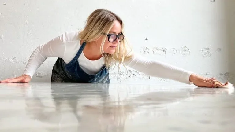 A woman wearing glasses and a blue apron is cleaning or polishing a large, flat surface, leaning forward with one hand extended and the other supporting her on the surface.