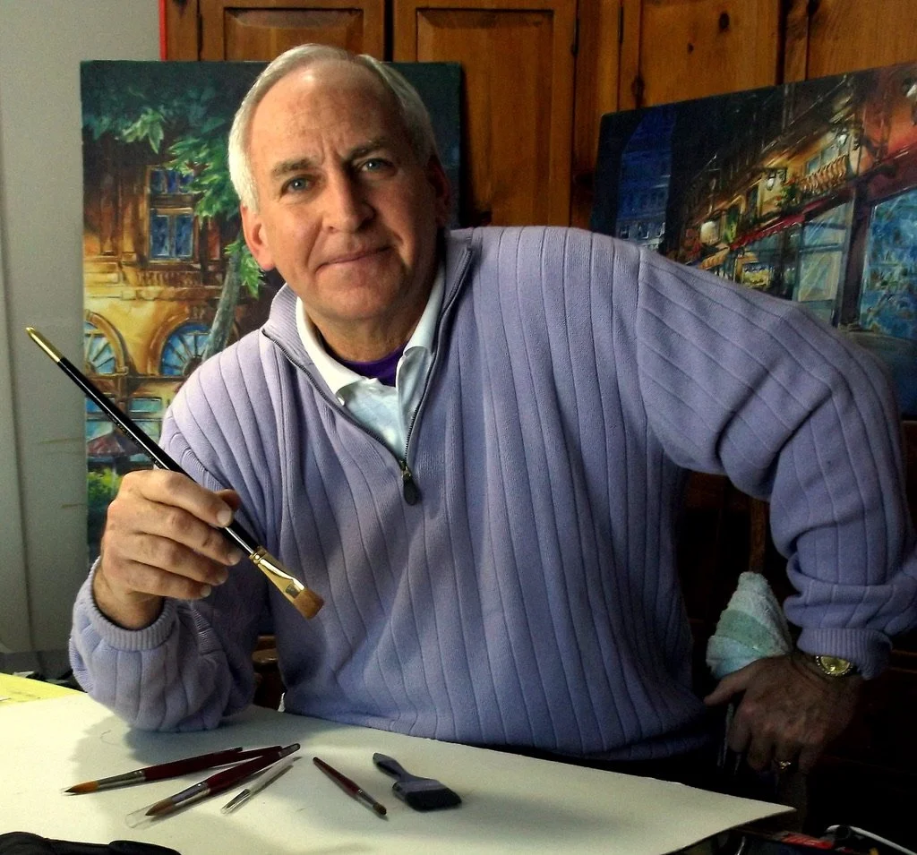 A man with gray hair, wearing a lavender sweater, holding a paintbrush, sitting at a table with paintbrushes, with paintings of cityscapes in the background.