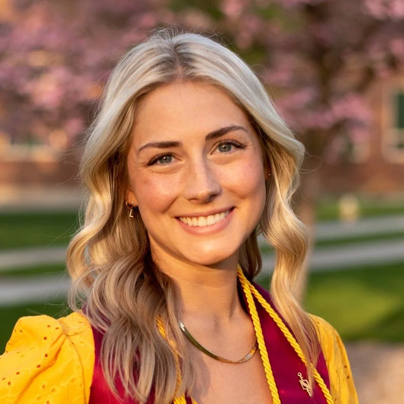A young woman with wavy blonde hair, wearing a maroon and yellow graduation gown and hood, smiling outdoors with pink blooming trees in the background.