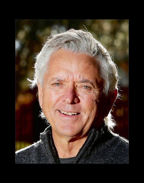 Close-up of an elderly man with gray hair, smiling outdoors with blurred trees in the background.