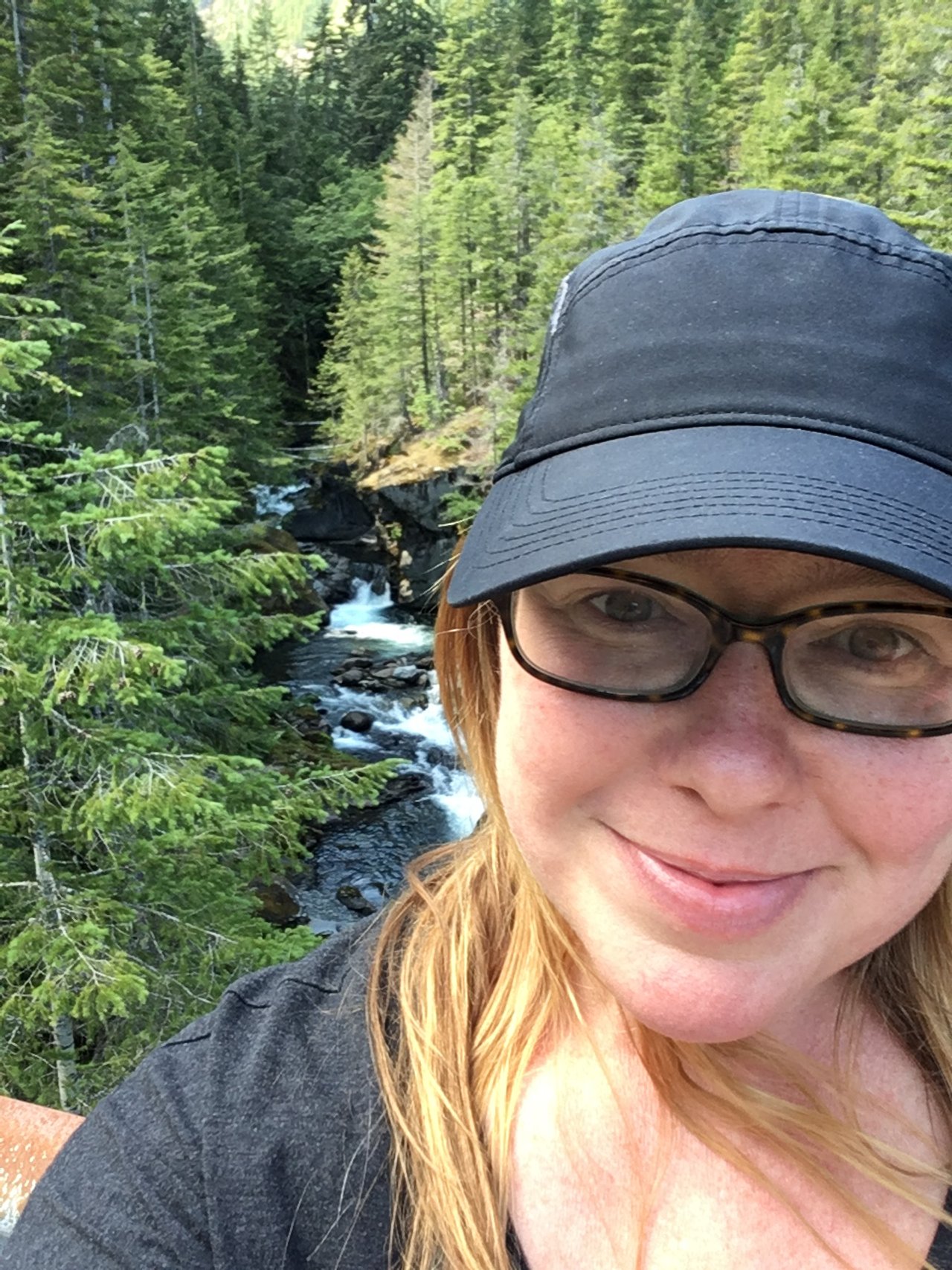 A woman wearing glasses and a black cap smiling outdoors with a forest and a flowing stream in the background.