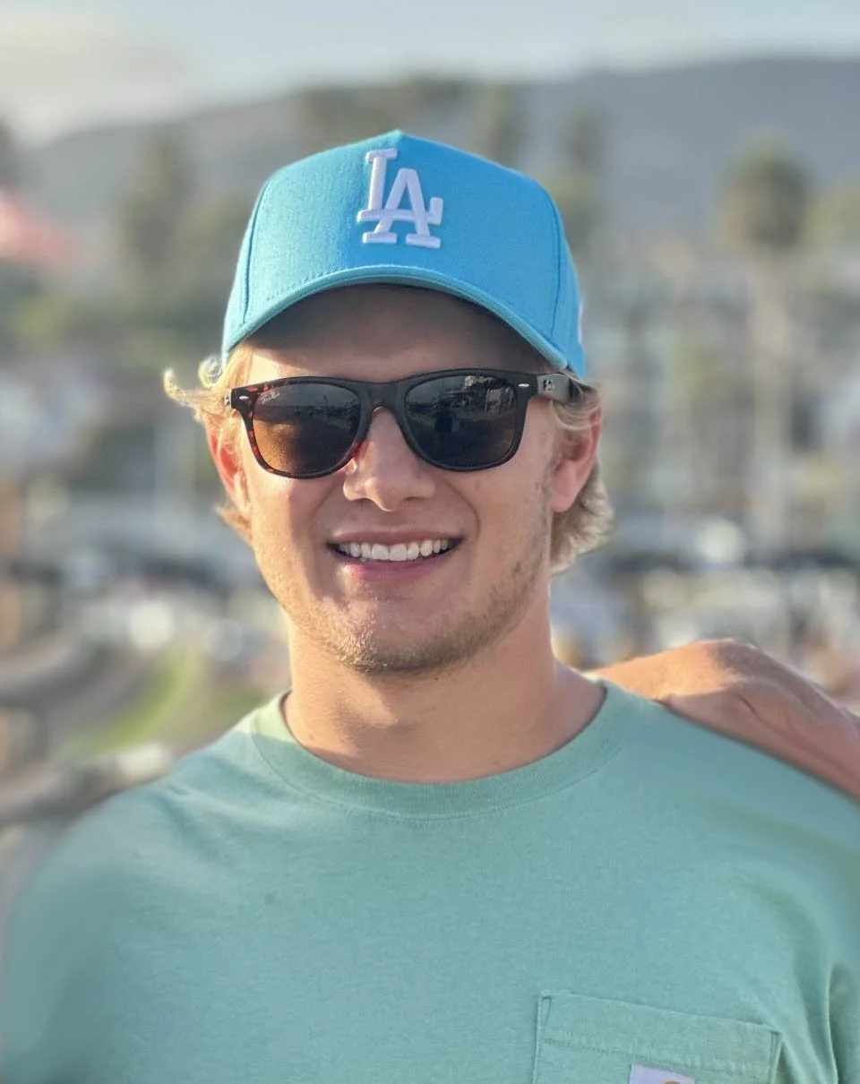 Young man smiling outdoors wearing a light blue Los Angeles Dodgers baseball cap, black sunglasses, and a mint green T-shirt.