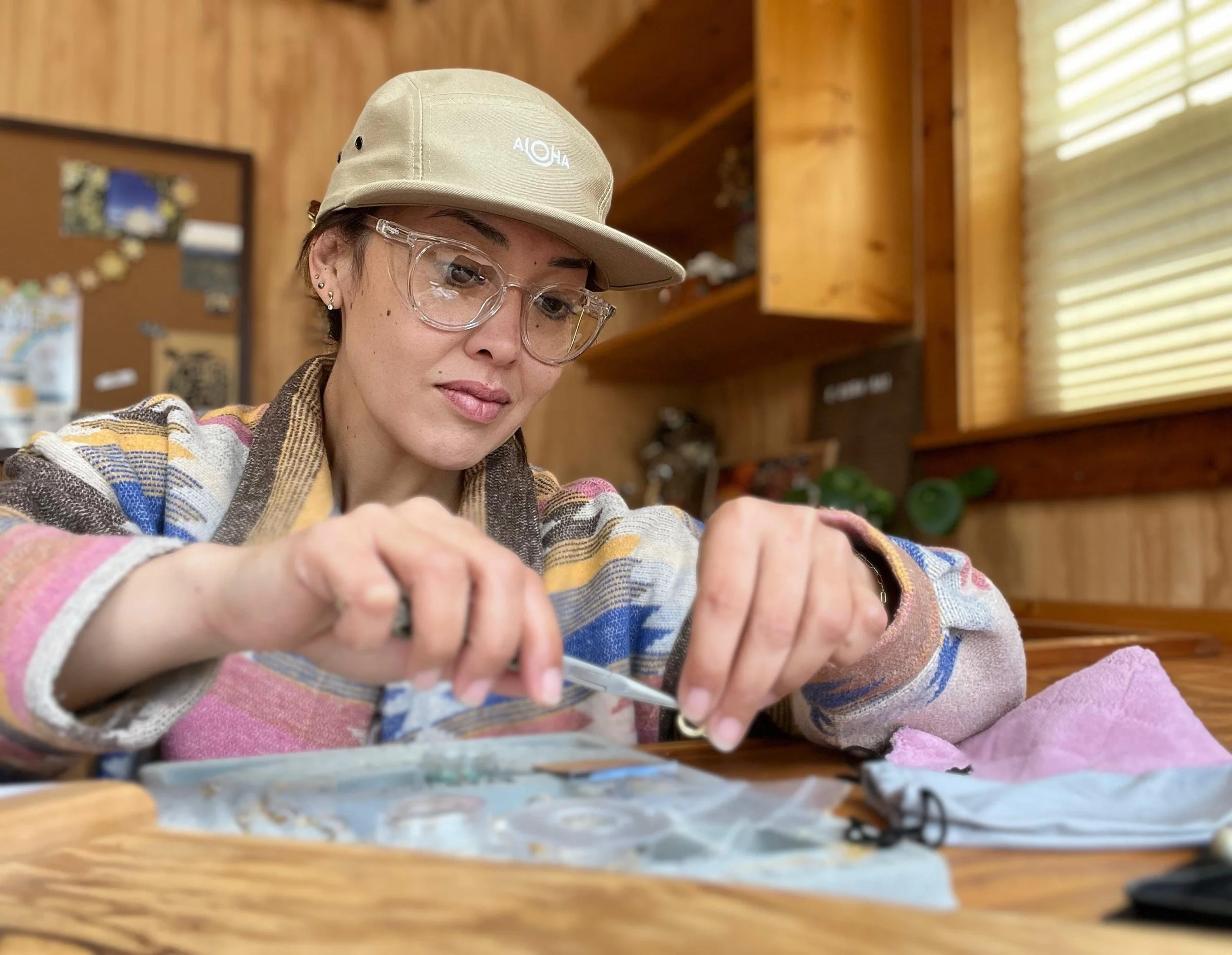 Young woman wearing glasses, a beige cap with white text, and a multicolored striped sweater, working at a wooden table in a cozy wood-paneled room with window blinds and a bulletin board in the background.