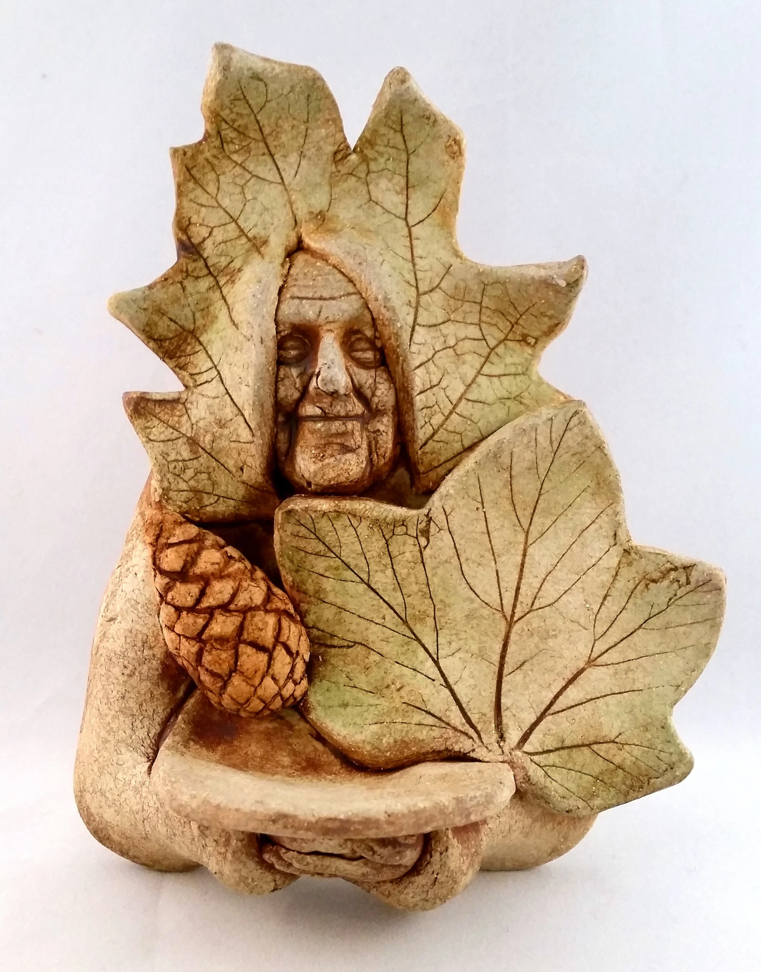 A small sculpture of an elderly woman with a serene expression, surrounded by autumn leaves and holding a pinecone.